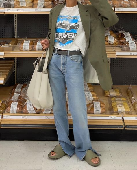 Person wearing a green blazer, graphic t-shirt, and jeans standing in a bread aisle of a supermarket with a white tote bag.