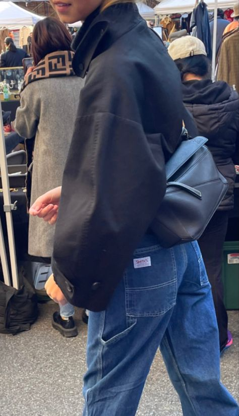 Person wearing a black jacket, blue jeans, and a black crossbody bag standing in a market.