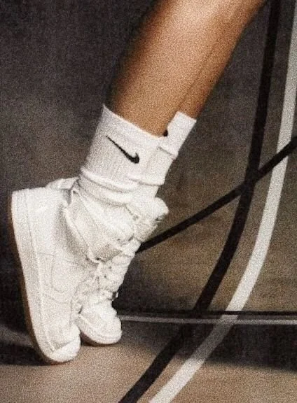 Person wearing white high-top sneakers and white socks with a black logo on a basketball court background.
