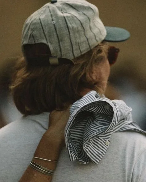 Back view of a person wearing a striped cap and sunglasses, holding a striped shirt over their shoulder.