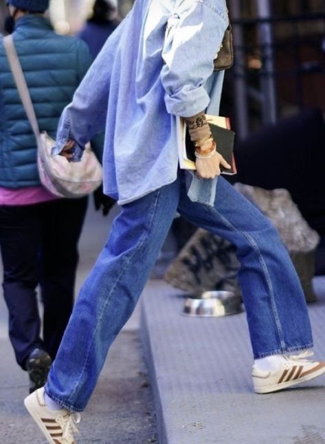 Person in denim outfit walking, wearing white sneakers with brown stripes, holding books.