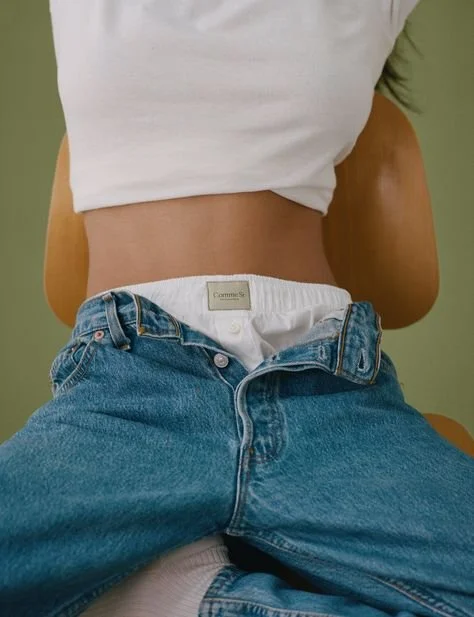 Person sitting with jeans unbuttoned and white tank top, showing waistband of underwear.