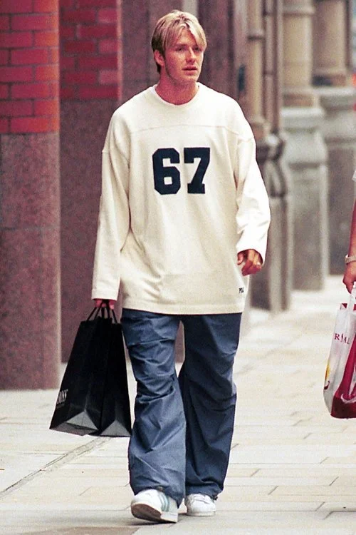 Person walking on a sidewalk wearing a white shirt with the number 67, blue pants, and holding shopping bags.