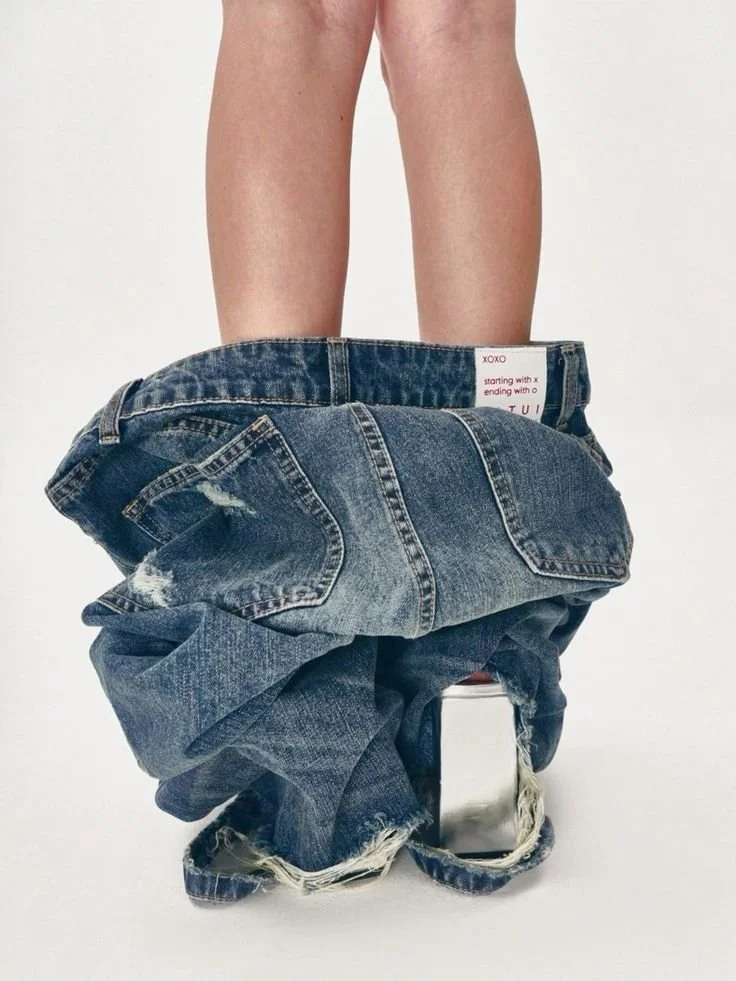 Person standing with jeans down around ankles, revealing legs and silver high-heeled shoes, studio background.