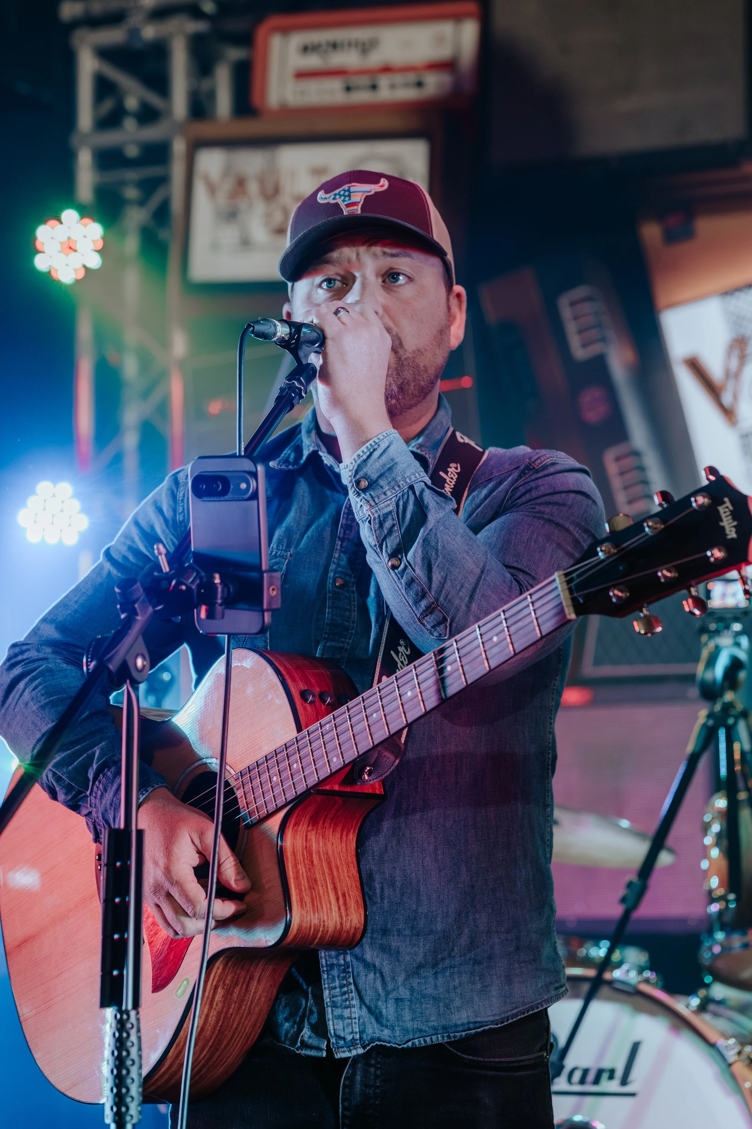 A man playing an acoustic guitar and singing into a microphone in a bar or pub setting, wearing a baseball cap and denim shirt.