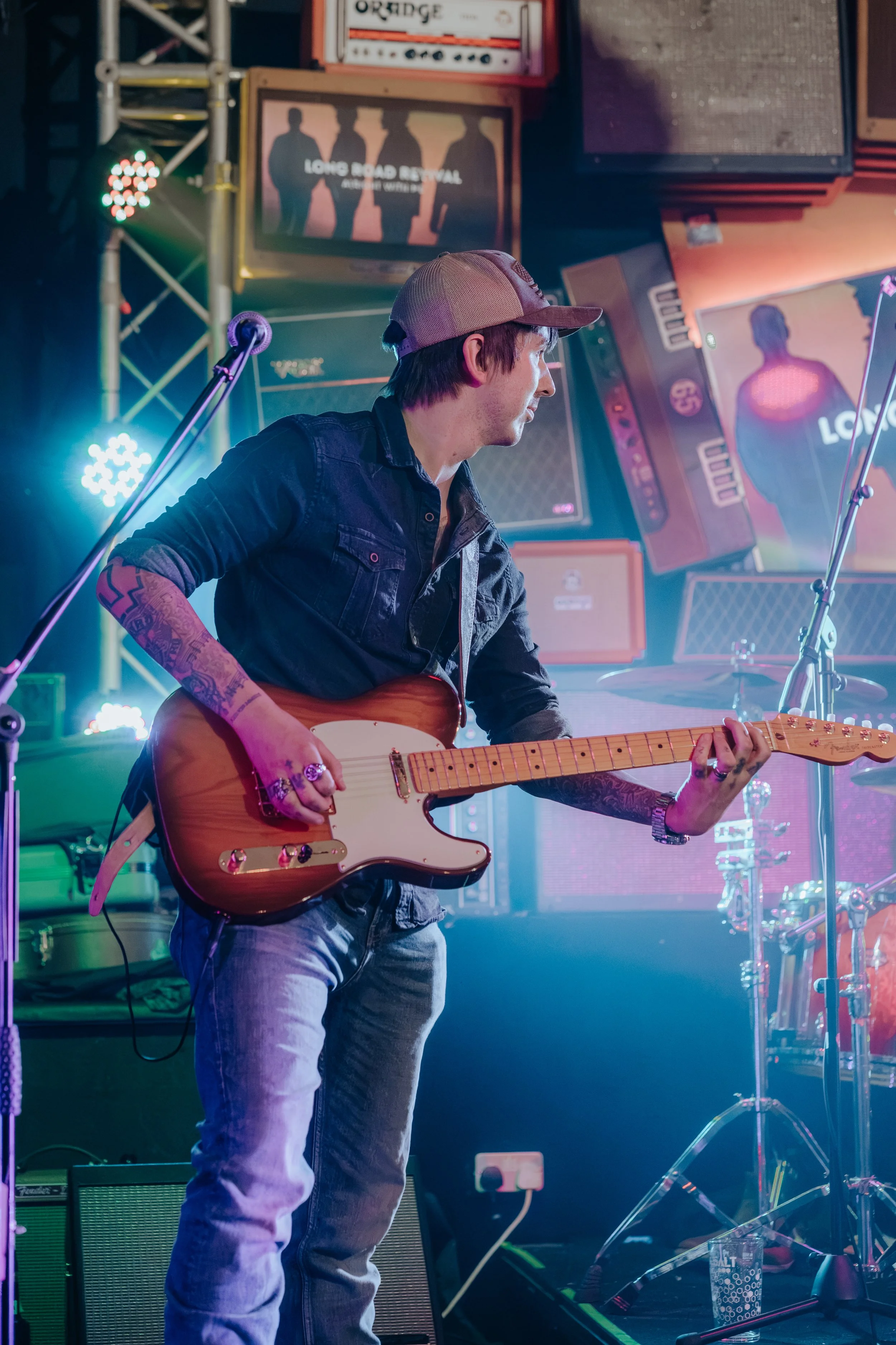 A man wearing a beige baseball cap, a dark denim shirt, and light jeans playing an electric guitar on stage with colorful lights and music equipment around him, including amplifiers, microphones, and speakers.
