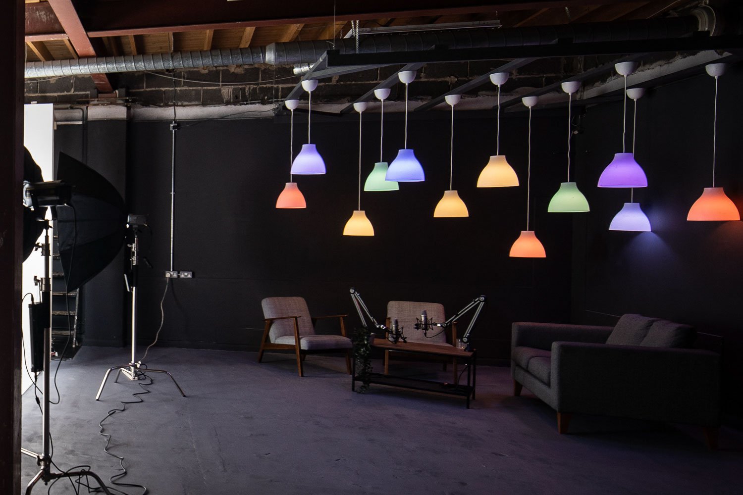 Indoor studio with colored pendant lights on black walls. Includes two armchairs, a sofa, a coffee table, and film or recording equipment.
