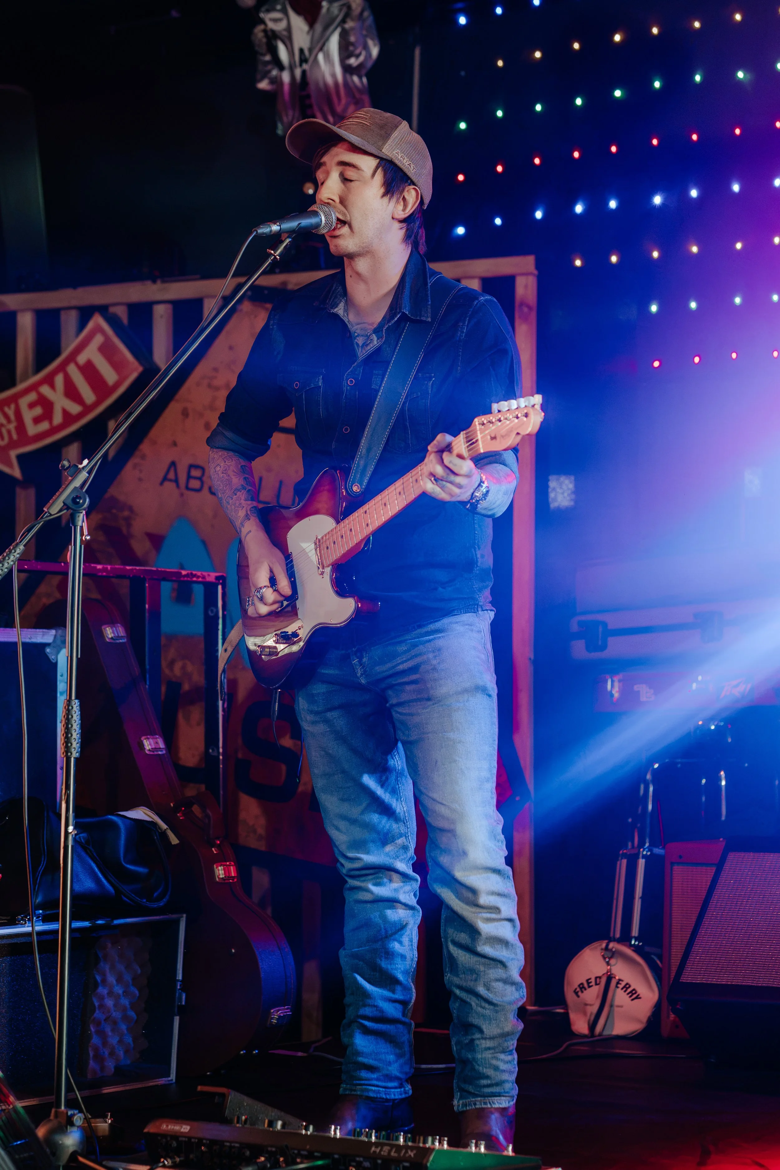 A male musician singing into a microphone, playing an electric guitar on a stage with colorful lights and a background sign.