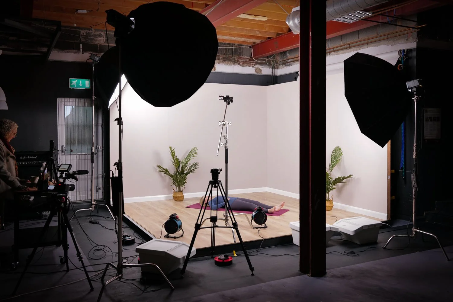 A woman is lying on her back on a yoga mat in a photography studio, surrounded by studio lights and equipment. There are two potted plants and a plain white wall in the background.