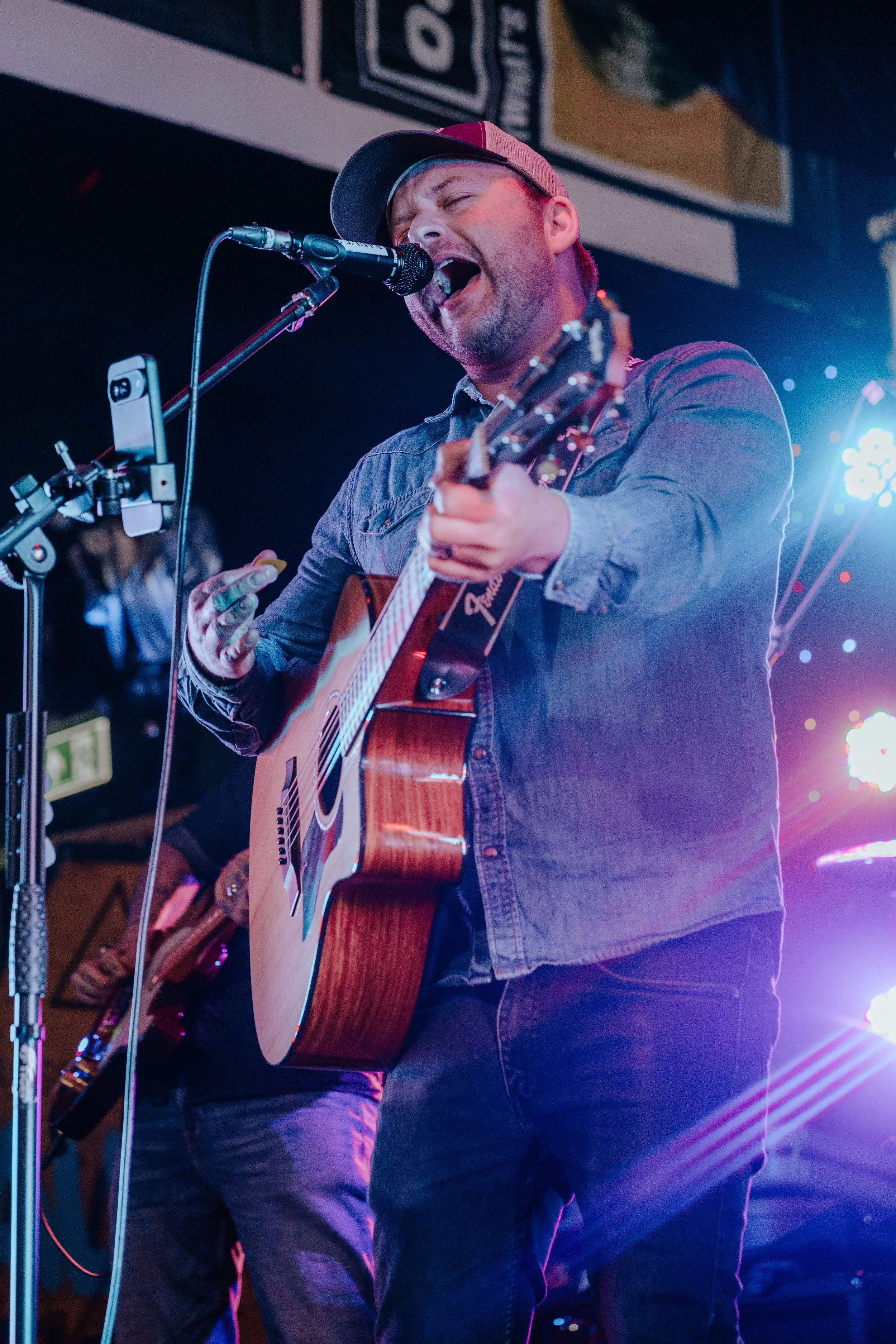 Male musician singing into a microphone while playing an acoustic guitar on stage under bright colorful lights.