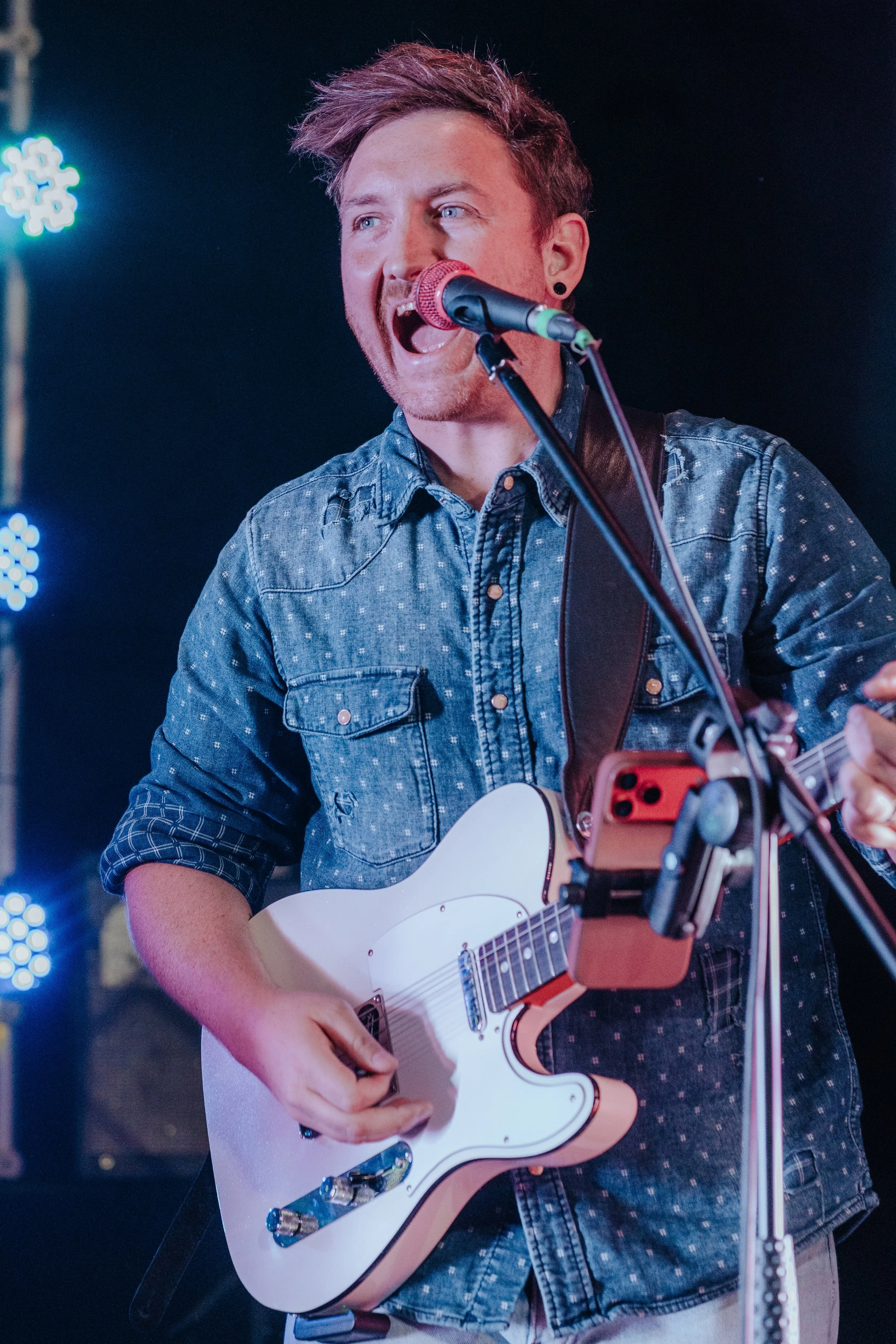 A young man with dyed hair and a beard, wearing a denim shirt, is singing and playing an electric guitar on stage with a microphone in front of him and blue stage lights in the background.