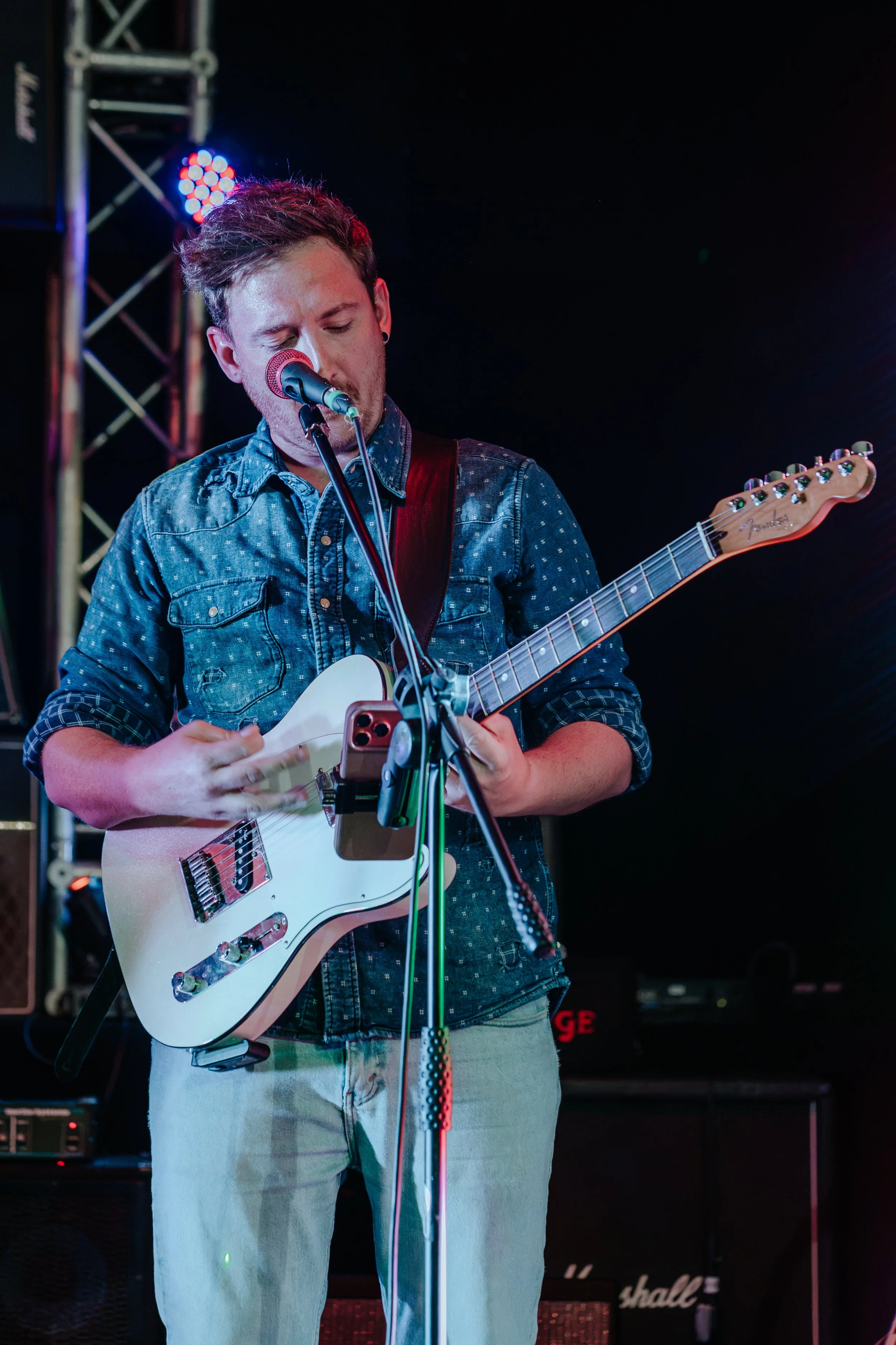 A young man performing on stage, singing into a microphone and playing an electric guitar, wearing a denim shirt and light-colored pants, with stage lights and equipment in the background.