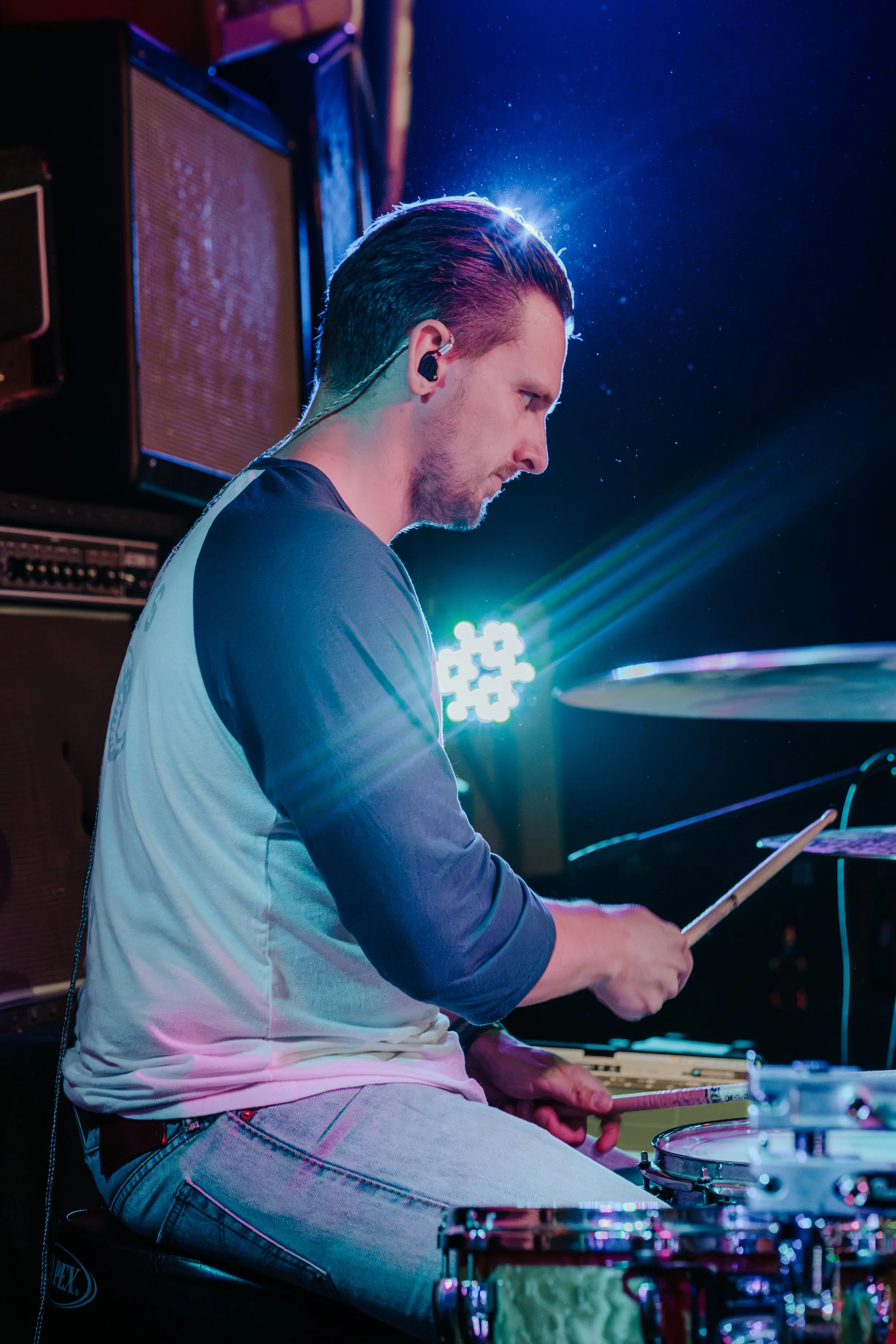 A male musician plays drums on stage, wearing a long-sleeved shirt and headphones, with colorful stage lighting and amplifiers in the background.