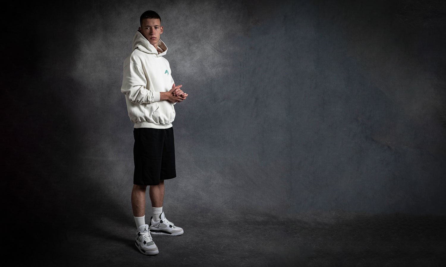 Young man standing against a dark gray textured background, wearing a white hoodie, black shorts, white socks, and gray and white sneakers, looking at the camera with hands clasped.