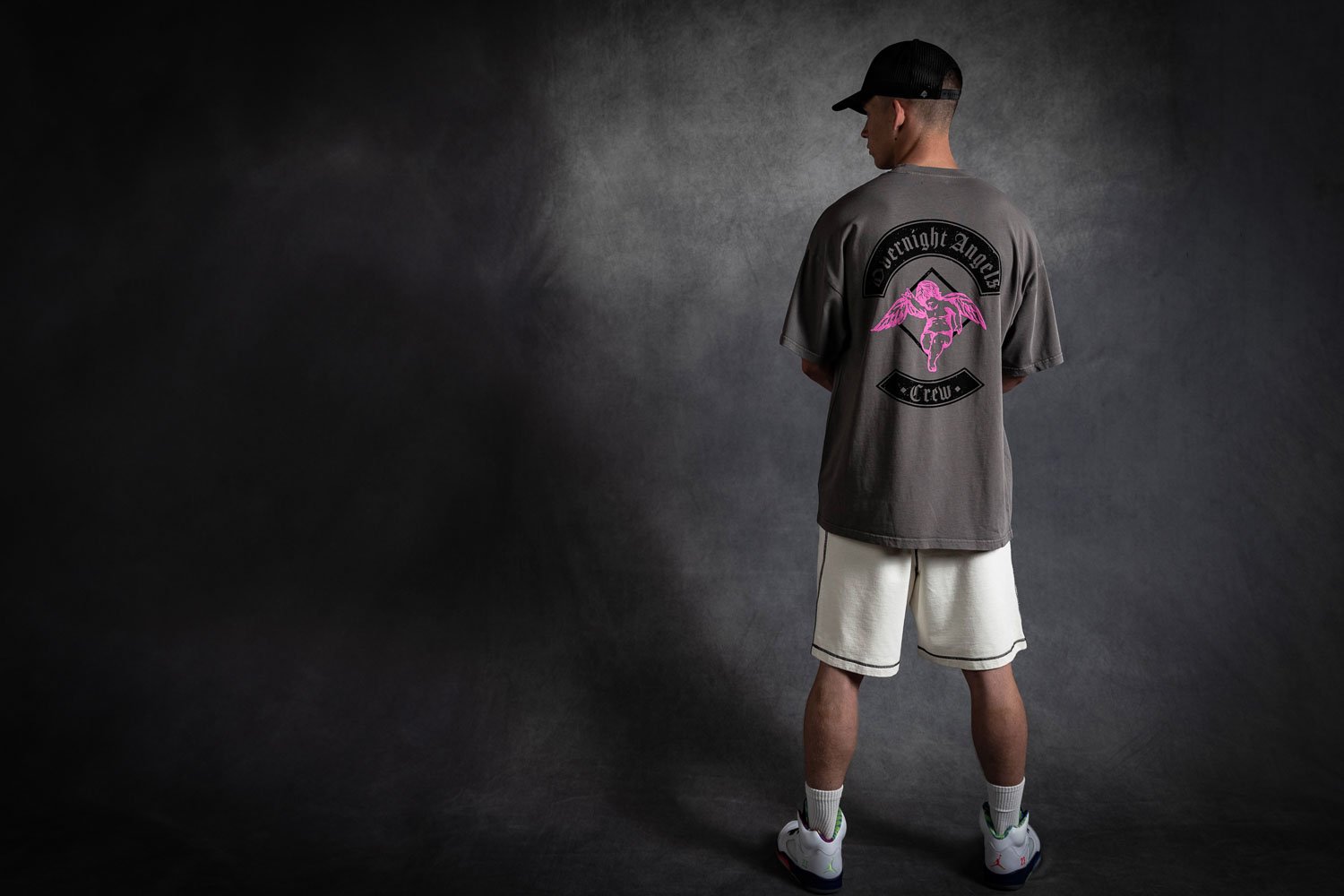 Person wearing a black baseball cap, a gray T-shirt with a pink graphic on the back, beige shorts, white sneakers, and white socks, standing against a dark, textured background.
