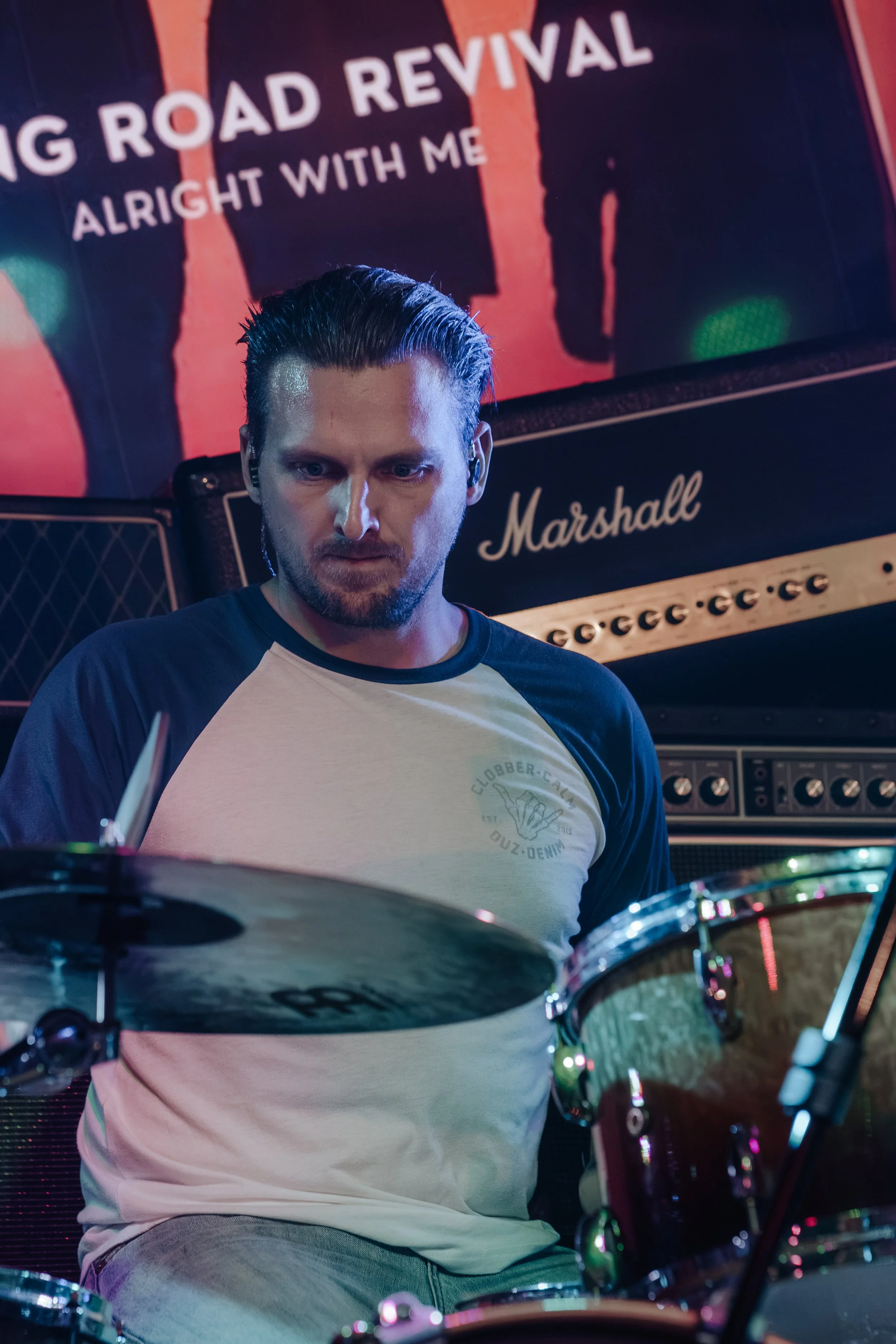 A man with dark hair and a beard playing drums in a music studio. He is wearing a beige and black baseball shirt and gray pants. A Marshall amplifier and a sign reading 'Big Road Revival Alright With Me' are visible in the background.