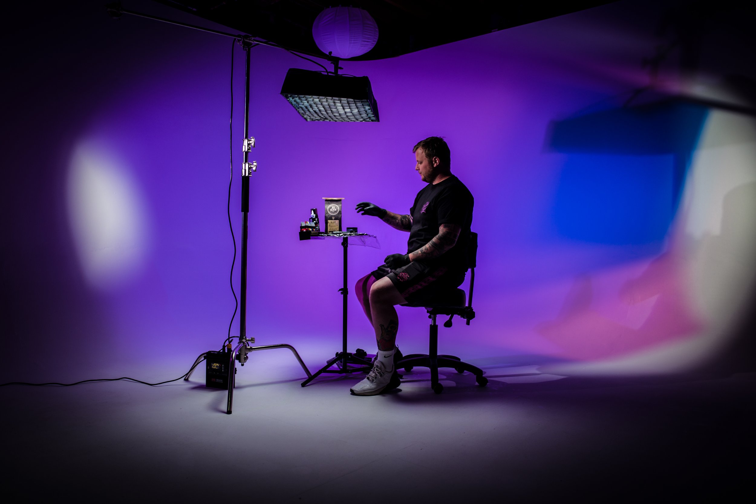 A man sitting on a rolling chair in a photography studio with purple lighting, wearing black gloves, shorts, and white sneakers, with photography equipment around him and a backdrop with a purple hue.
