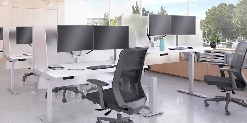 Modern office workspace with multiple desks, black monitors, ergonomic chairs, and large windows showing outside greenery.