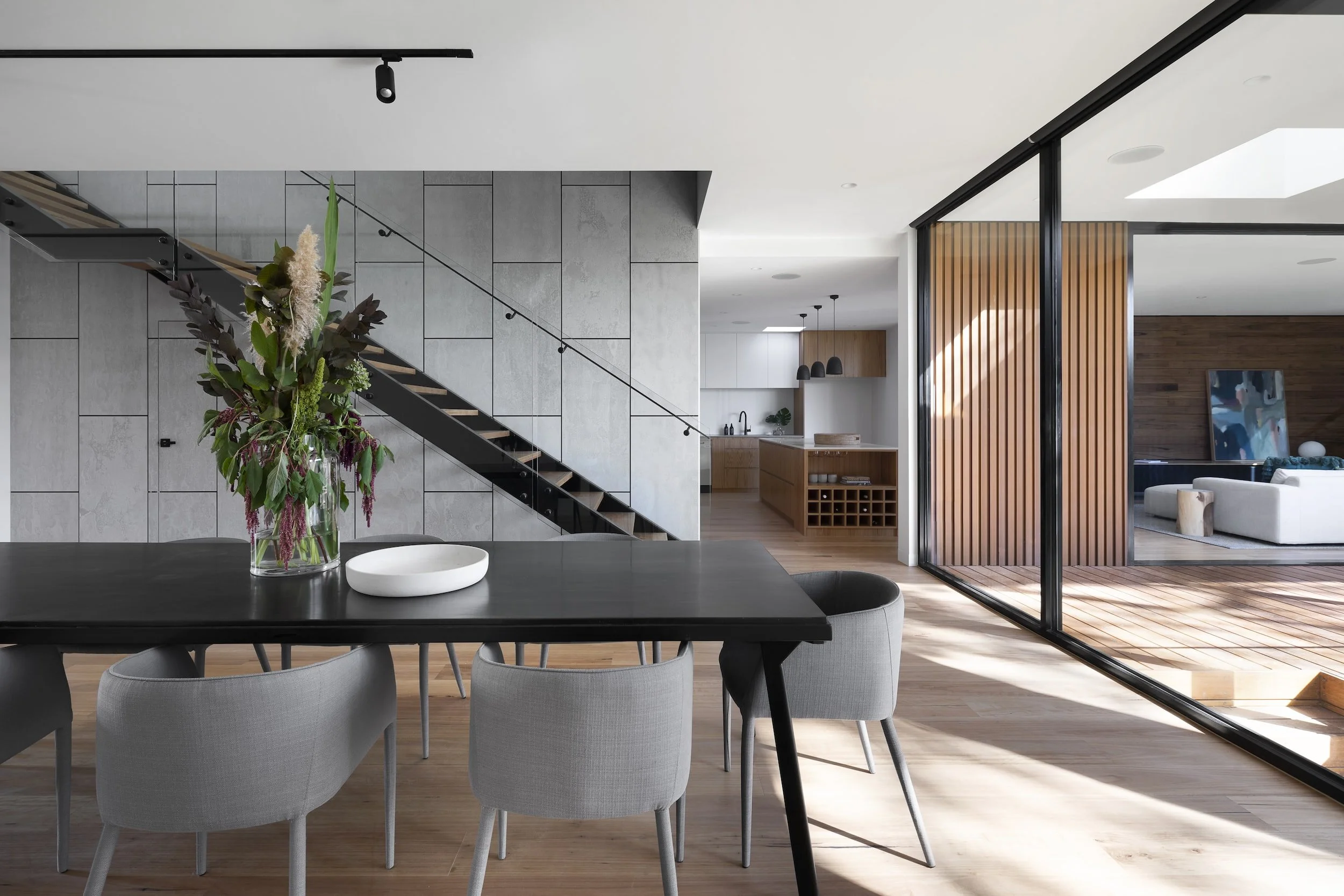 Modern dining room with a black table, gray chairs, and a large floral arrangement. Adjacent is a staircase with glass railing, and in the background a kitchen with pendant lights and wooden cabinetry. Floor-to-ceiling windows provide natural light.