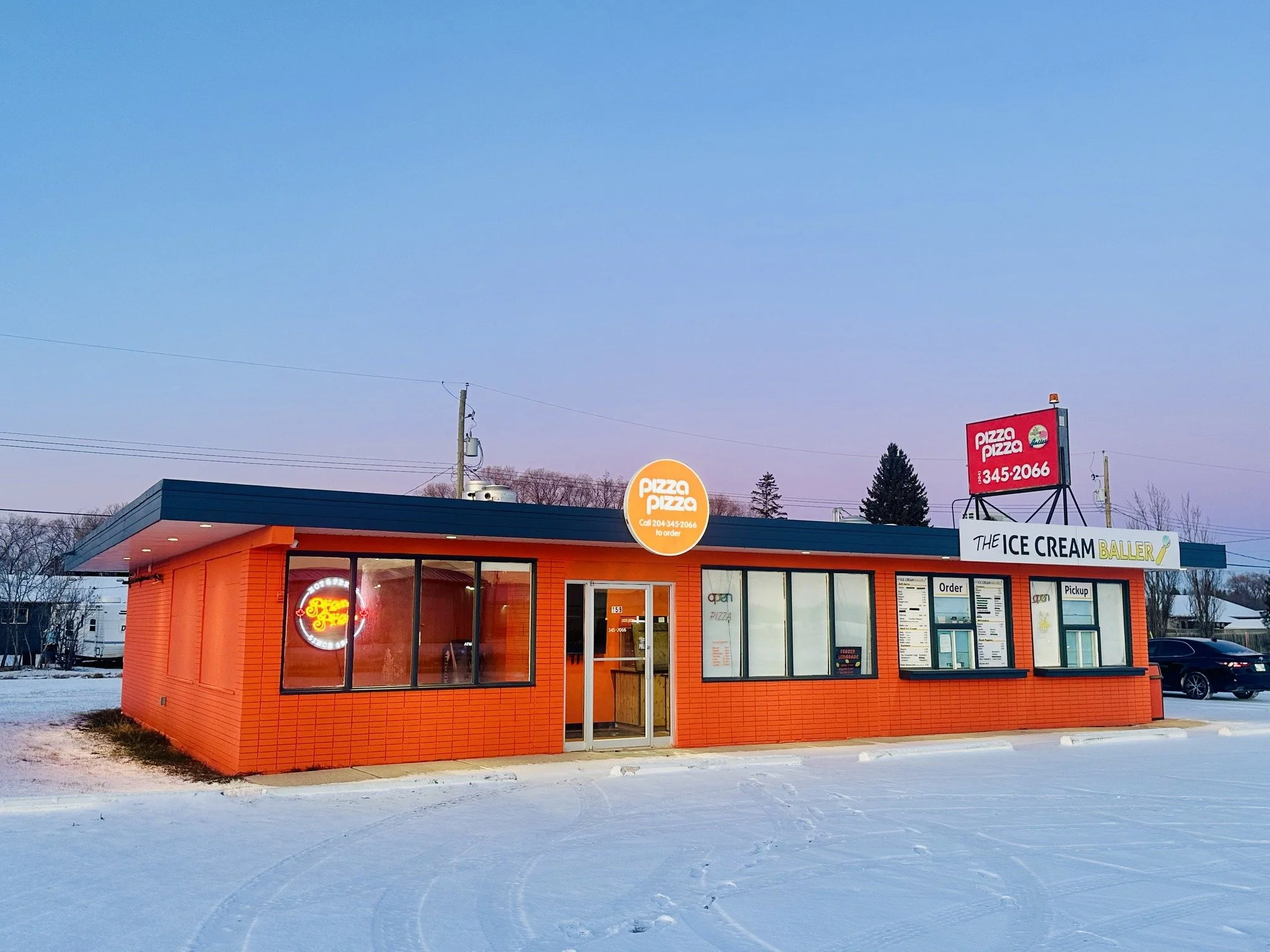 Orange brick restaurant building with large glass windows, a side door, and signs for pizza and ice cream, situated on snow-covered ground with a clear evening sky.