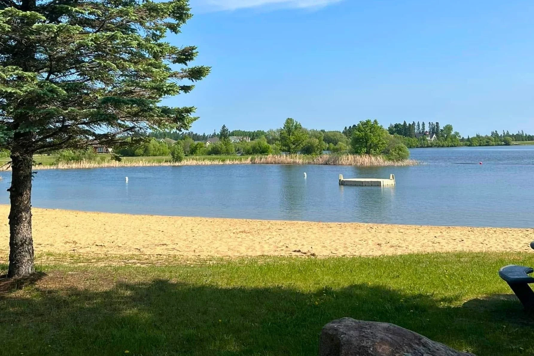 Showcasing St-Georges Beach, a gorgeous Winnipeg River beach with a floating swim platform, treed area, and mix of sand and grass.