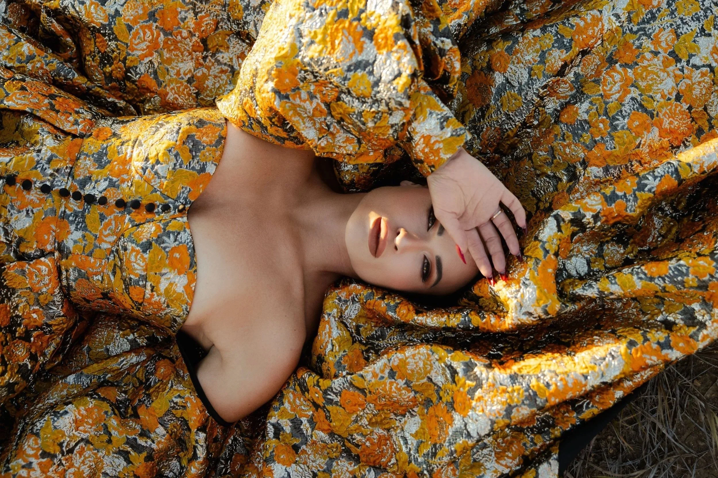 Woman lying down outdoors, wearing a floral robe with gold and orange patterns, covering part of her face with her hand, with land visible in the background.