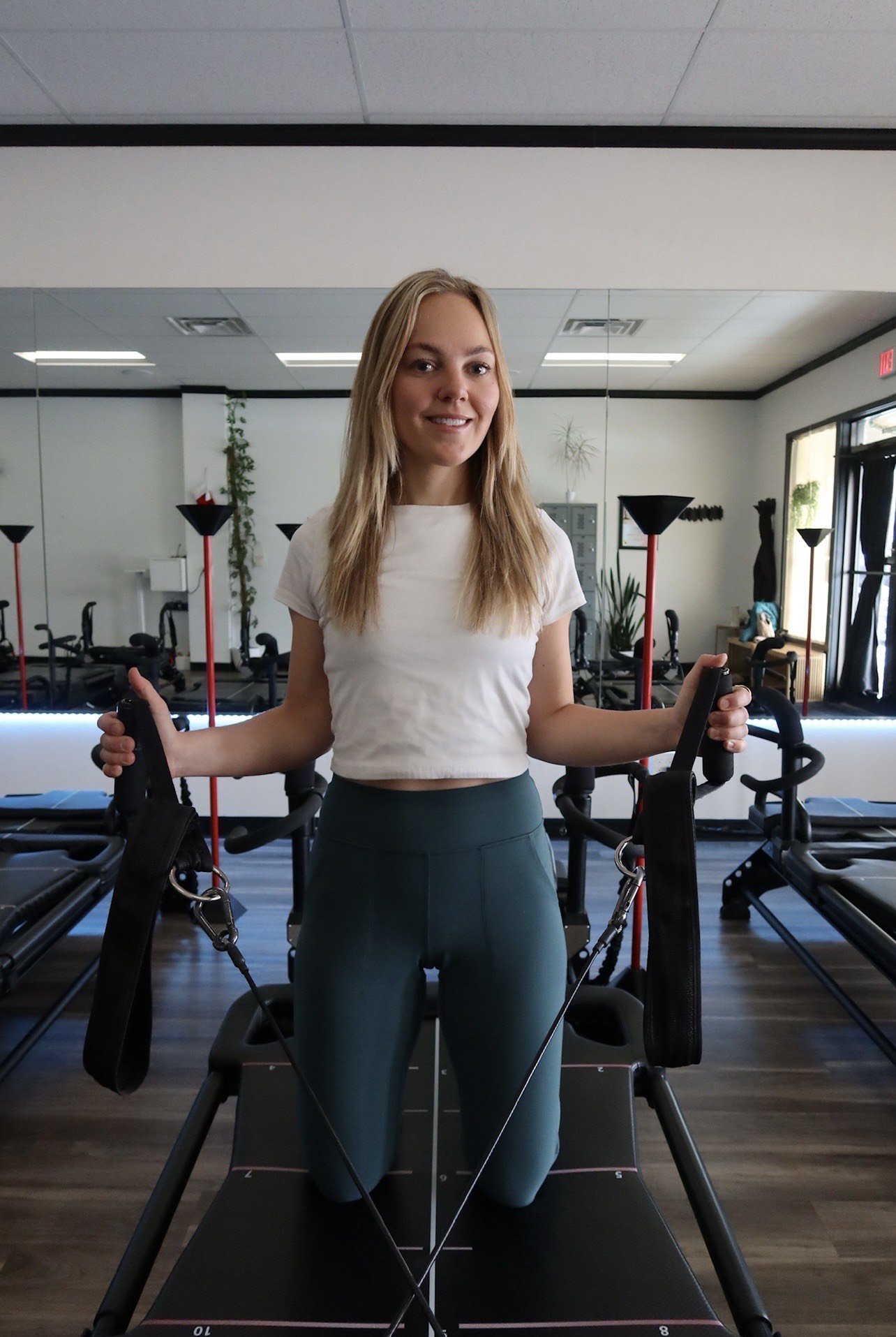 A woman in workout clothes kneeling on a reformer pilates machine in a gym, holding the straps in both hands, with mirror reflections and gym equipment in the background.