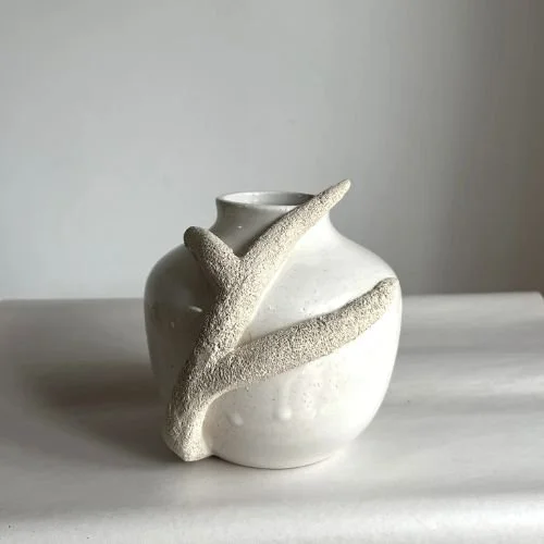 White ceramic vase with a textured, branch-like handle on a light surface against a plain background.
