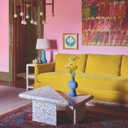 Living room with a yellow sofa, pink walls, and abstract art. There is a white side table with a blue lamp and a framed picture. A pink ceiling light fixture is visible.