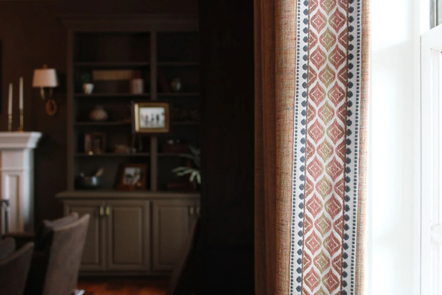 Den with earthy brown color-drenched walls and built-in bookshelves, layered drapery fabrics, natural plants and greenery, and a mix of new and antique furniture creating a warm, inviting space.