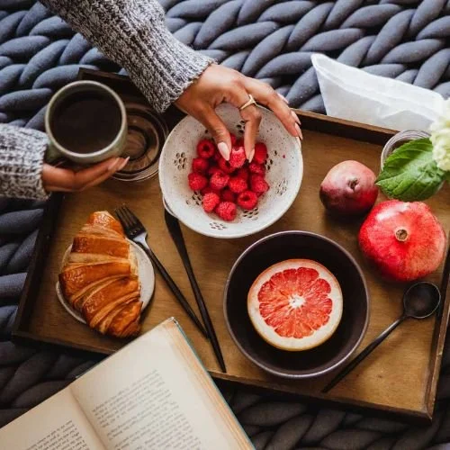 A wooden tray with a cup of coffee, a bowl of raspberries, a grapefruit half, a pomegranate, and a green leaf with a tissue and an open book, on a textured gray blanket.