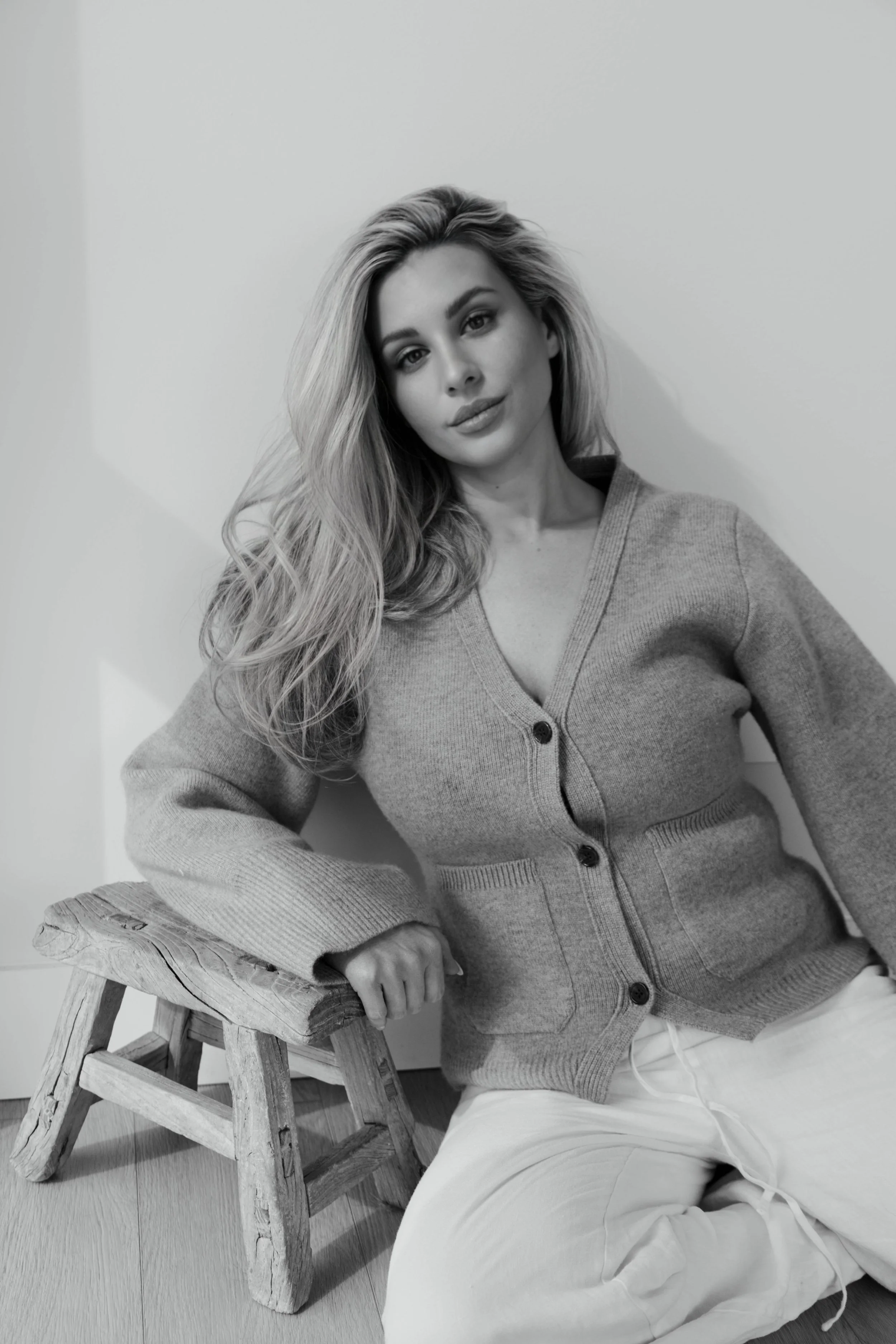 Black and white photo of a woman with long wavy hair sitting on a wooden stool, wearing a cardigan and a skirt, against a plain wall background.