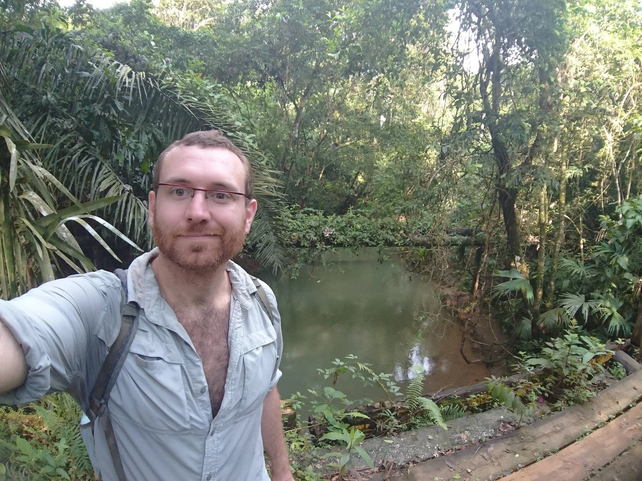 A man with glasses and a beard taking a selfie in a lush, green jungle near a river.