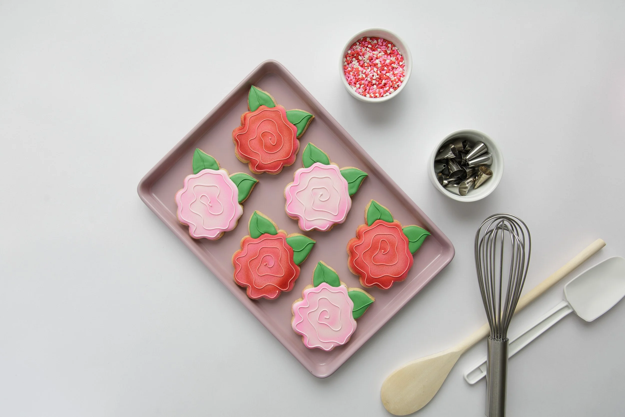 Product photography flatlay showing rose-shaped cookies styled with simple tools and a ramekin to create clean, cohesive imagery for small business marketing.