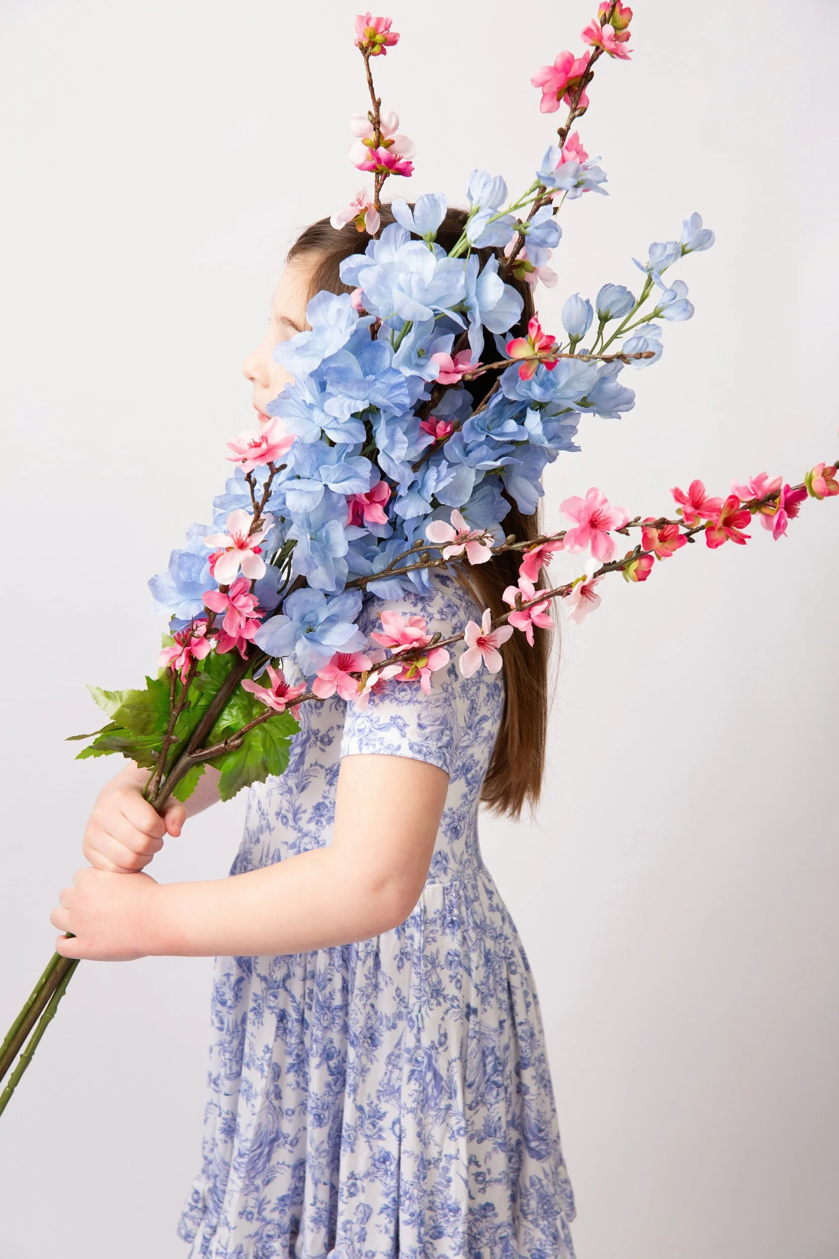 Little girl with long brown hair wearing a blue floral dress and holding a bouquet of long-stem spring flowers in a bright white studio in Skippack, PA.