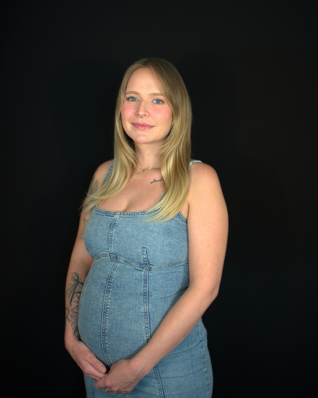 Portrait of a pregnant woman with long blonde hair, wearing a denim dress with floral straps, standing against a black background.