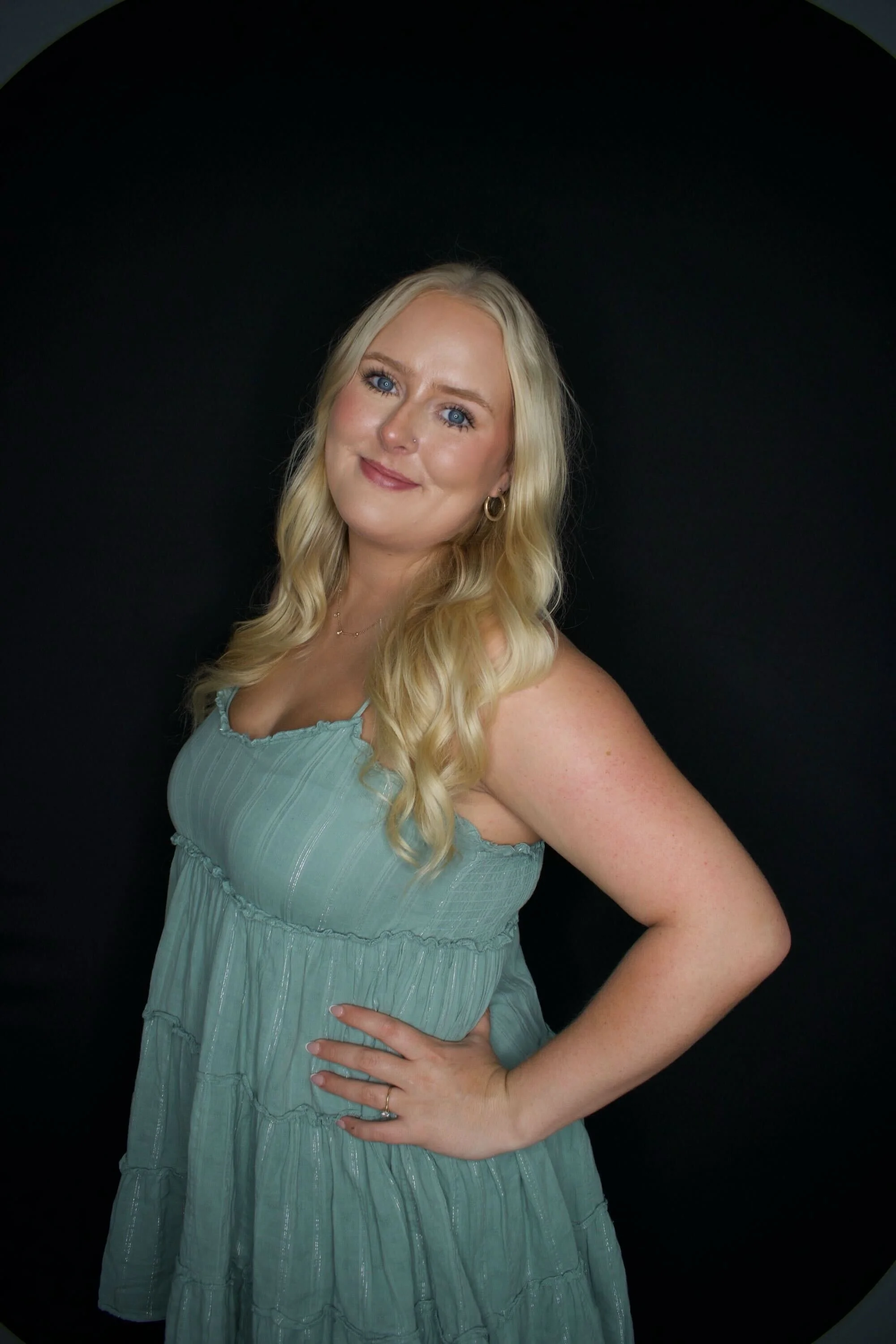 Woman with long blonde hair wearing a white top and black leather pants, smiling with hands in pockets, against a dark background.