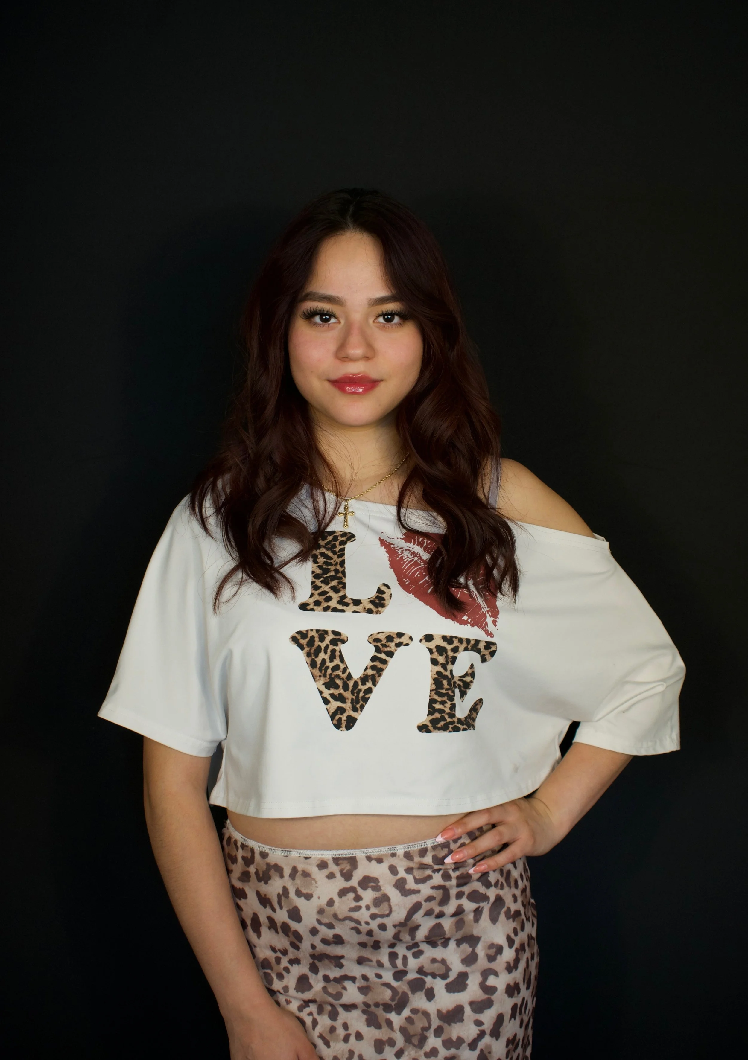Young woman with long wavy dark hair standing against a black background, wearing a white cropped T-shirt with the word 'LOVE' in leopard print letters and a red lipstick kiss mark, and a matching leopard print skirt.