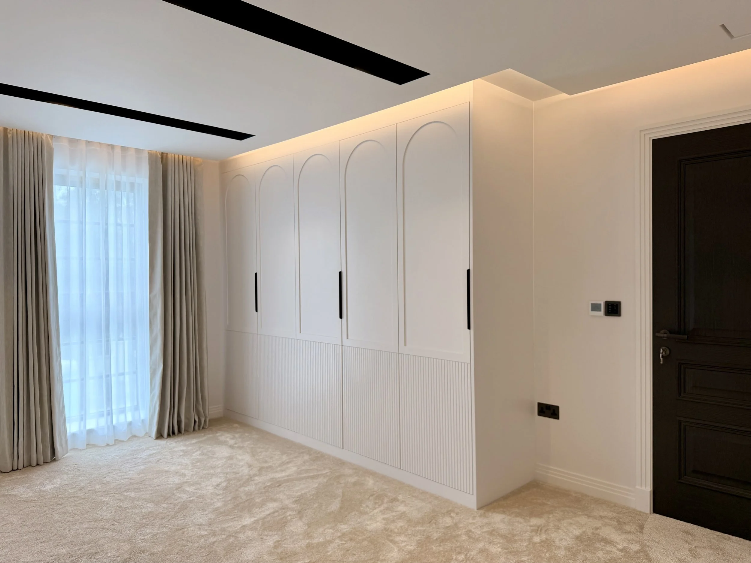 Handcrafted fitted wardrobes with elegant arched panel detailing and fluted lower sections, designed to complement the architecture of a luxury bedroom. Integrated storage solutions and carefully considered proportions create a timeless bespoke furni