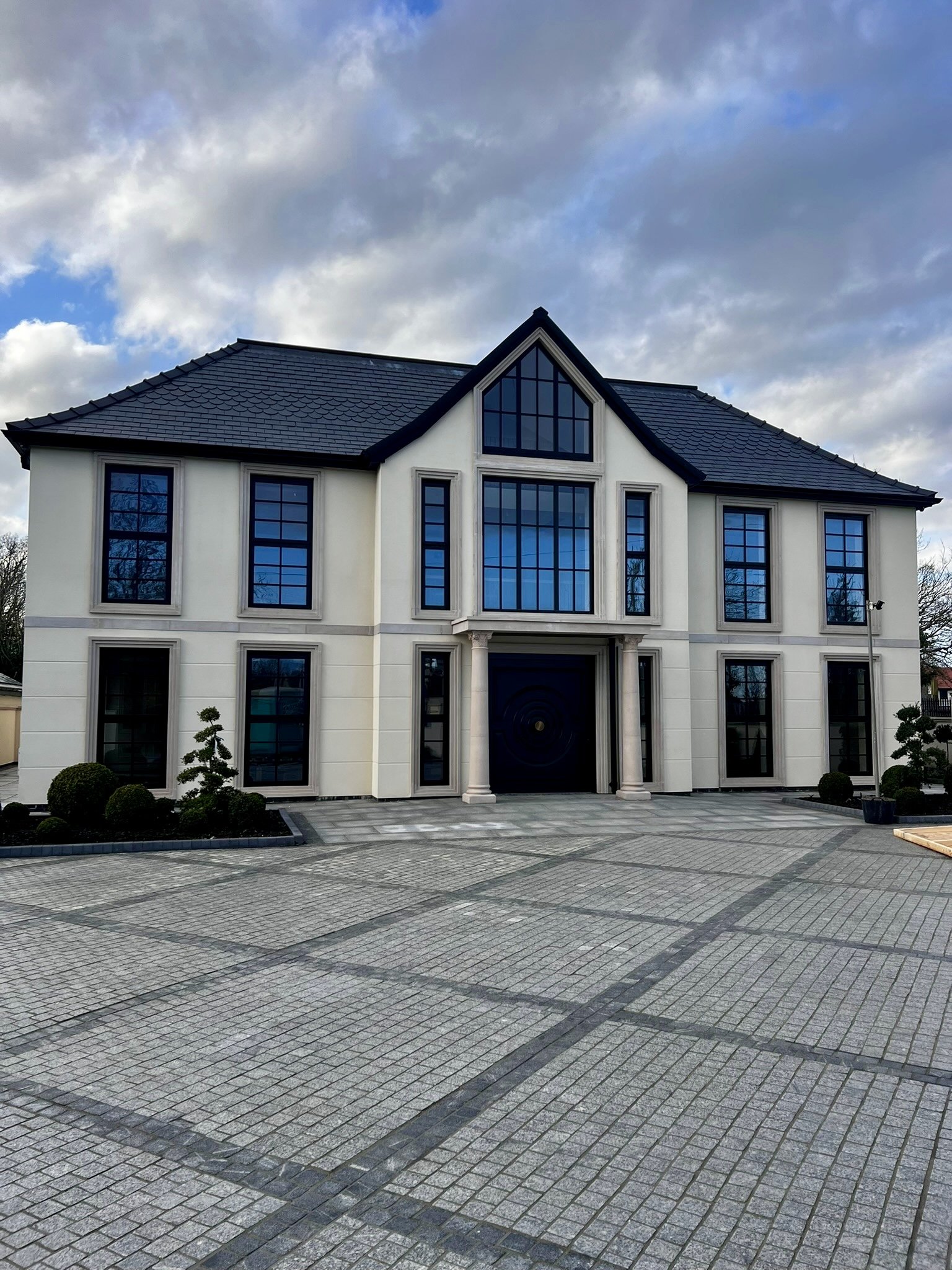 Modern luxury mansion with cream exterior walls, large black-framed windows, and a dark slate roof, featuring a grand central entrance with columns and an expansive paved driveway under a cloudy sky.