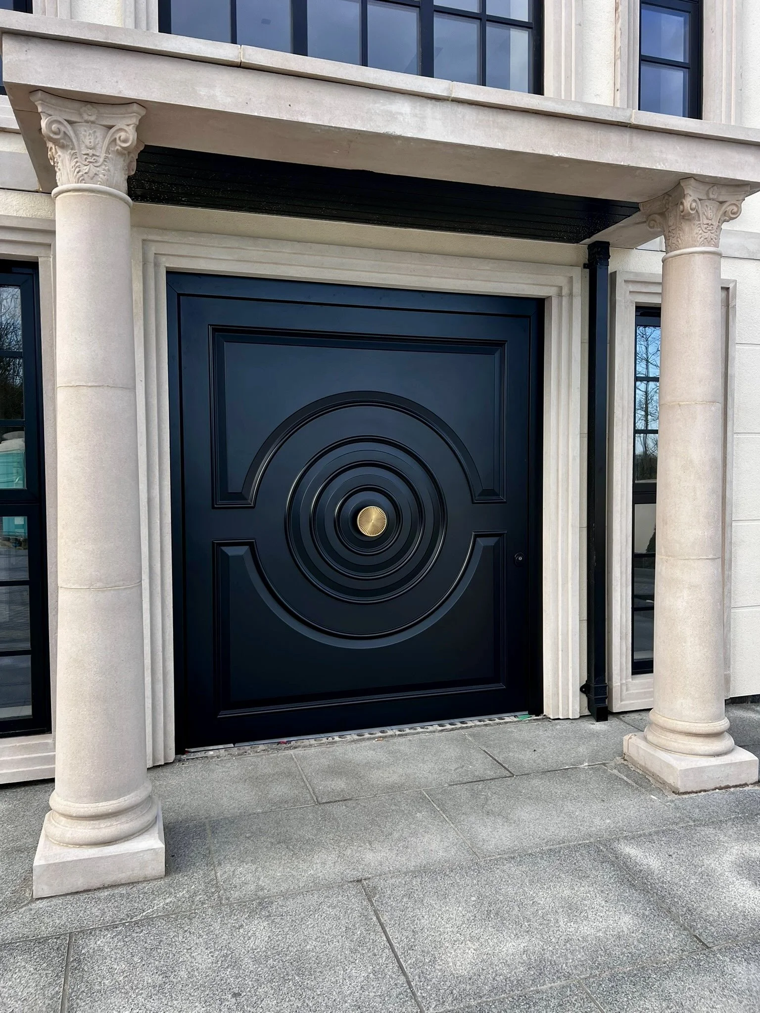 Luxury black front entrance door with circular geometric detailing, framed by elegant stone columns and modern architectural trim on a high-end contemporary mansion.