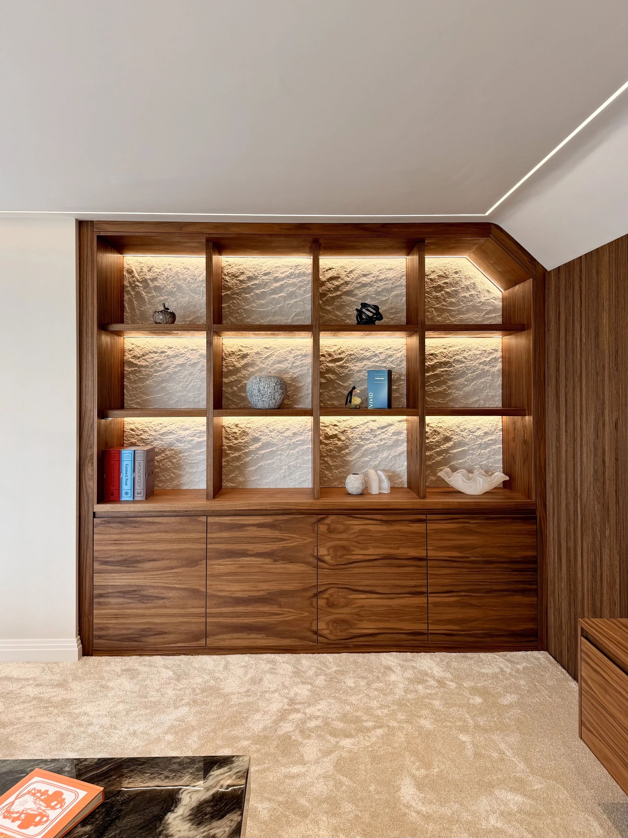 Custom walnut cabinetry with illuminated display shelving and textured feature panels, handcrafted to create a striking focal point within a luxury interior. Integrated LED lighting highlights decorative objects while concealed storage provides pract