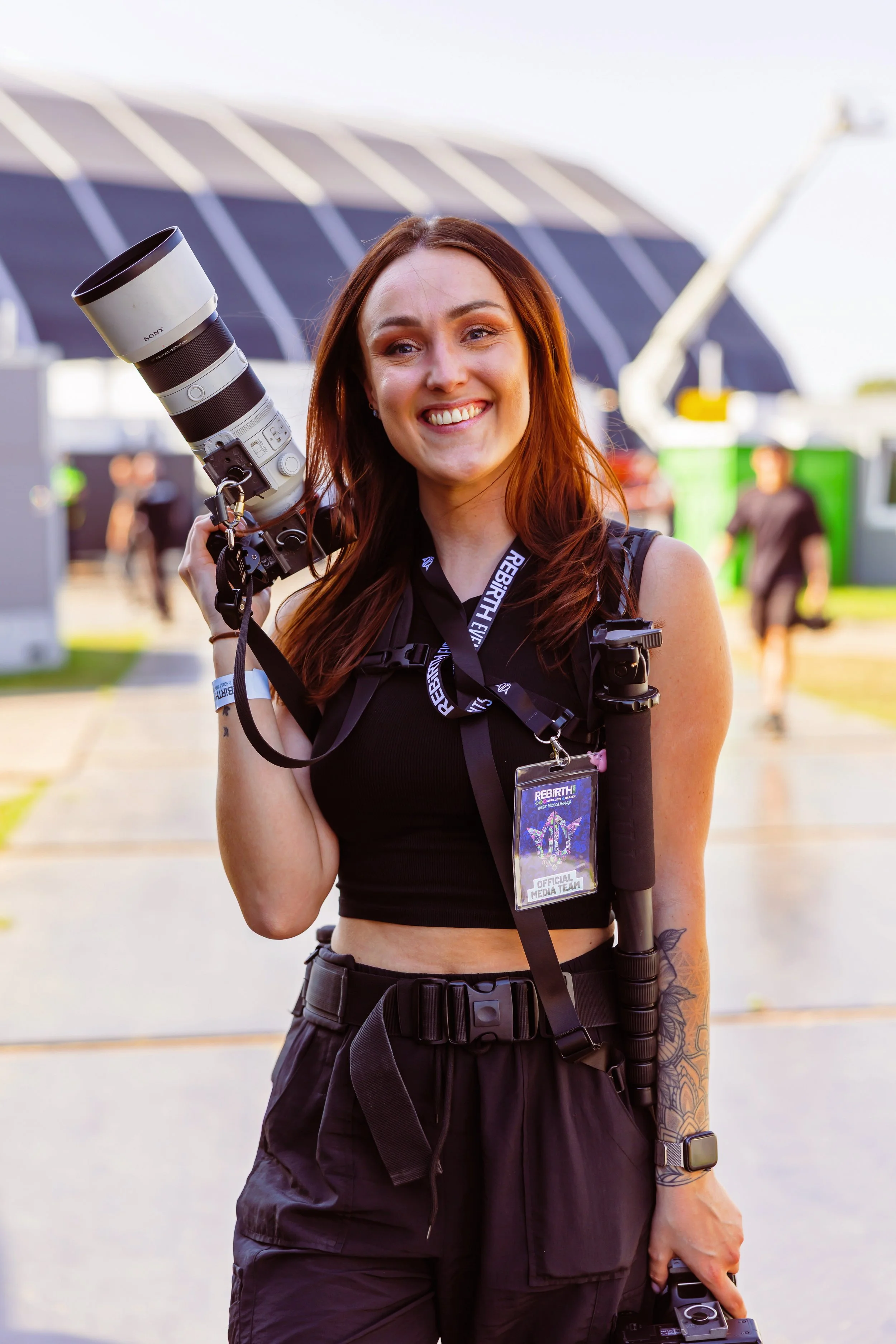Vrouw met een glimlach, met een grote camera op haar schouder, buiten bij een evenement.