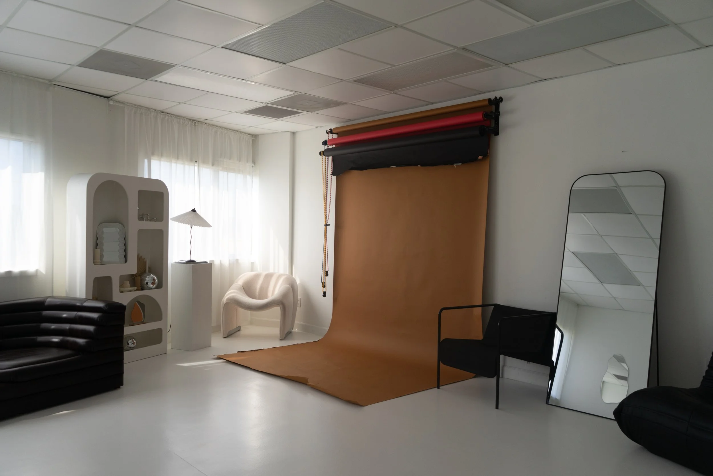 Photography studio with a brown backdrop, white shelf with decor, black sofa, cream armchair, black chair, and tall mirror.