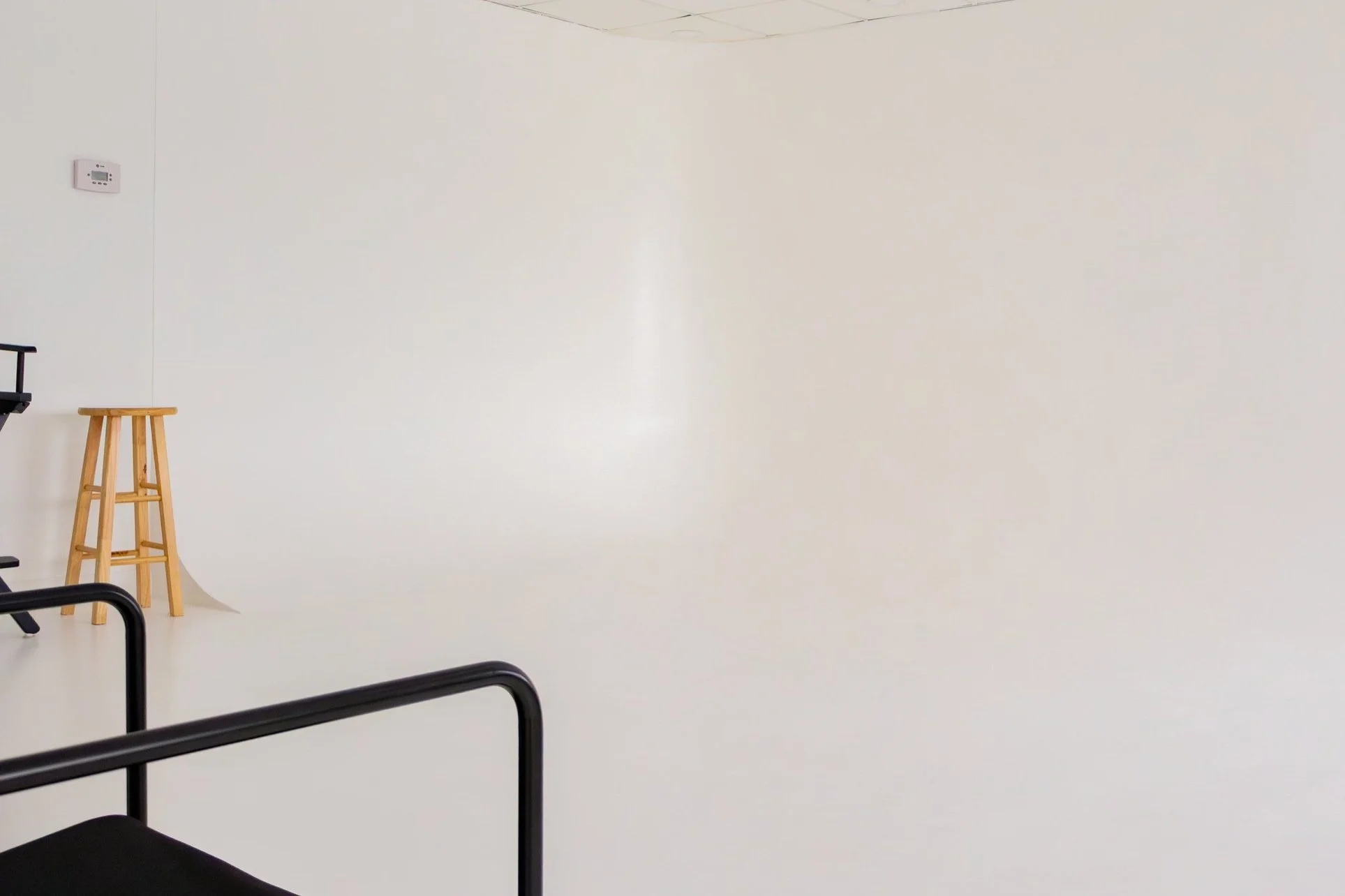 Empty studio with white walls, black metal chair, wooden stool, and a thermostat on the wall.