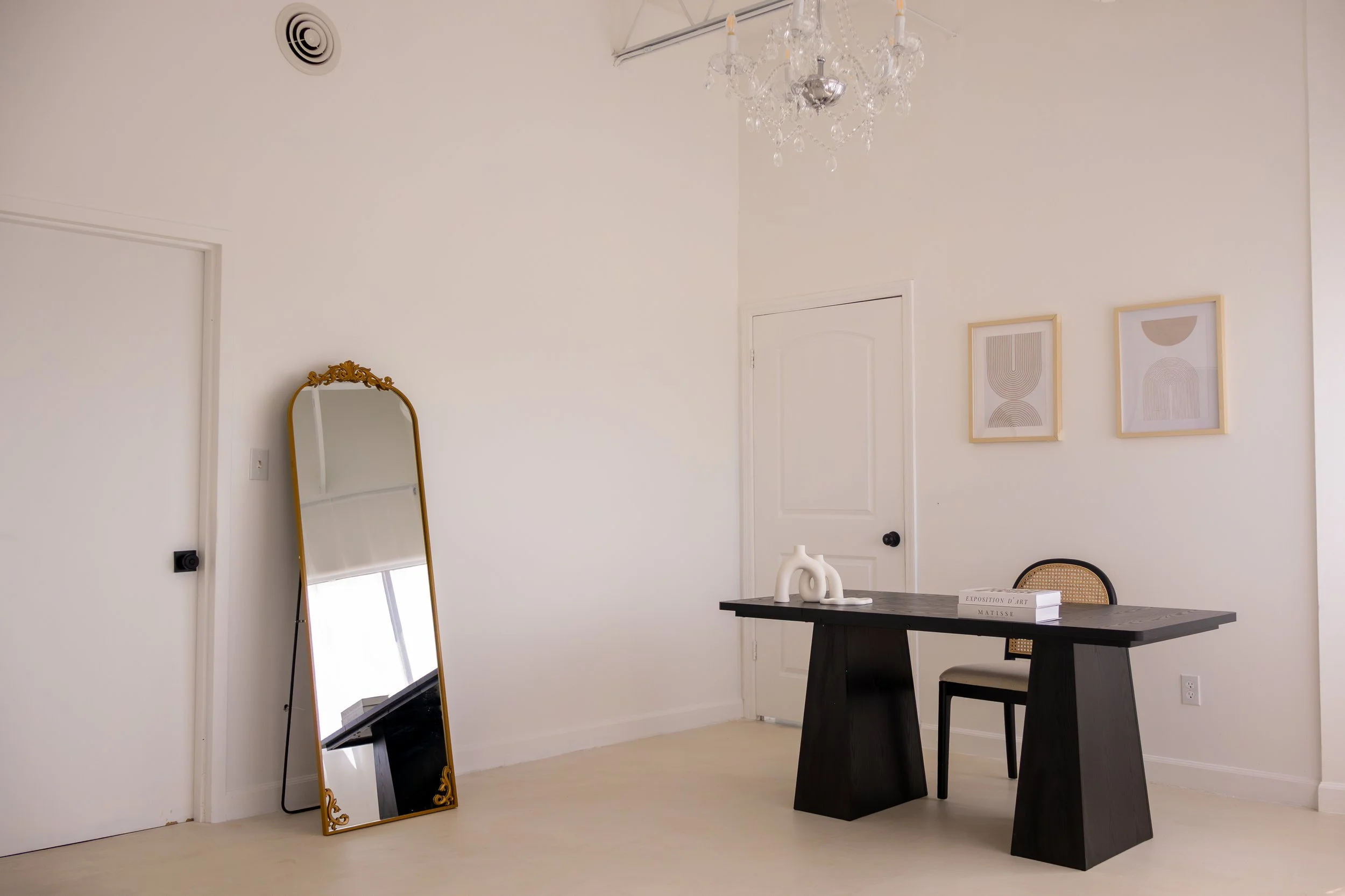 Minimalist interior with a large decorative mirror, a black table adorned with white sculptures and a book, a rattan-backed chair, framed abstract art on the wall, and a chandelier hanging from the ceiling.