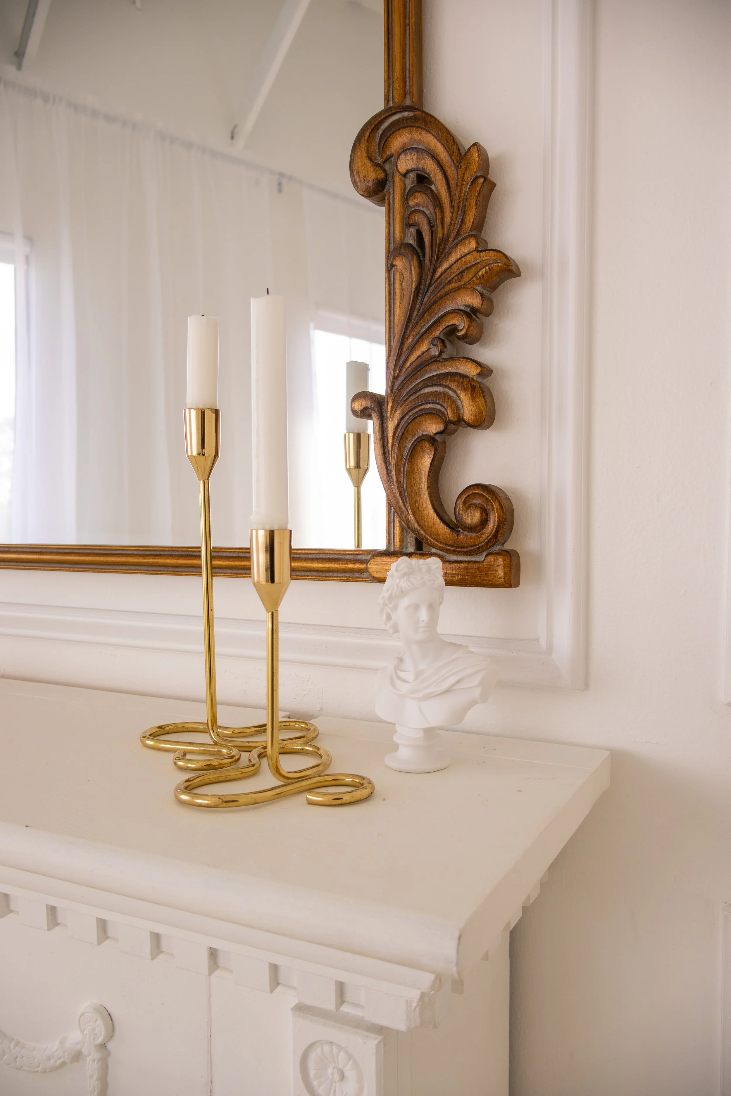 Decorative white mantel with a gold candle holder, a white bust sculpture, and a large wooden-framed mirror in a bright room.