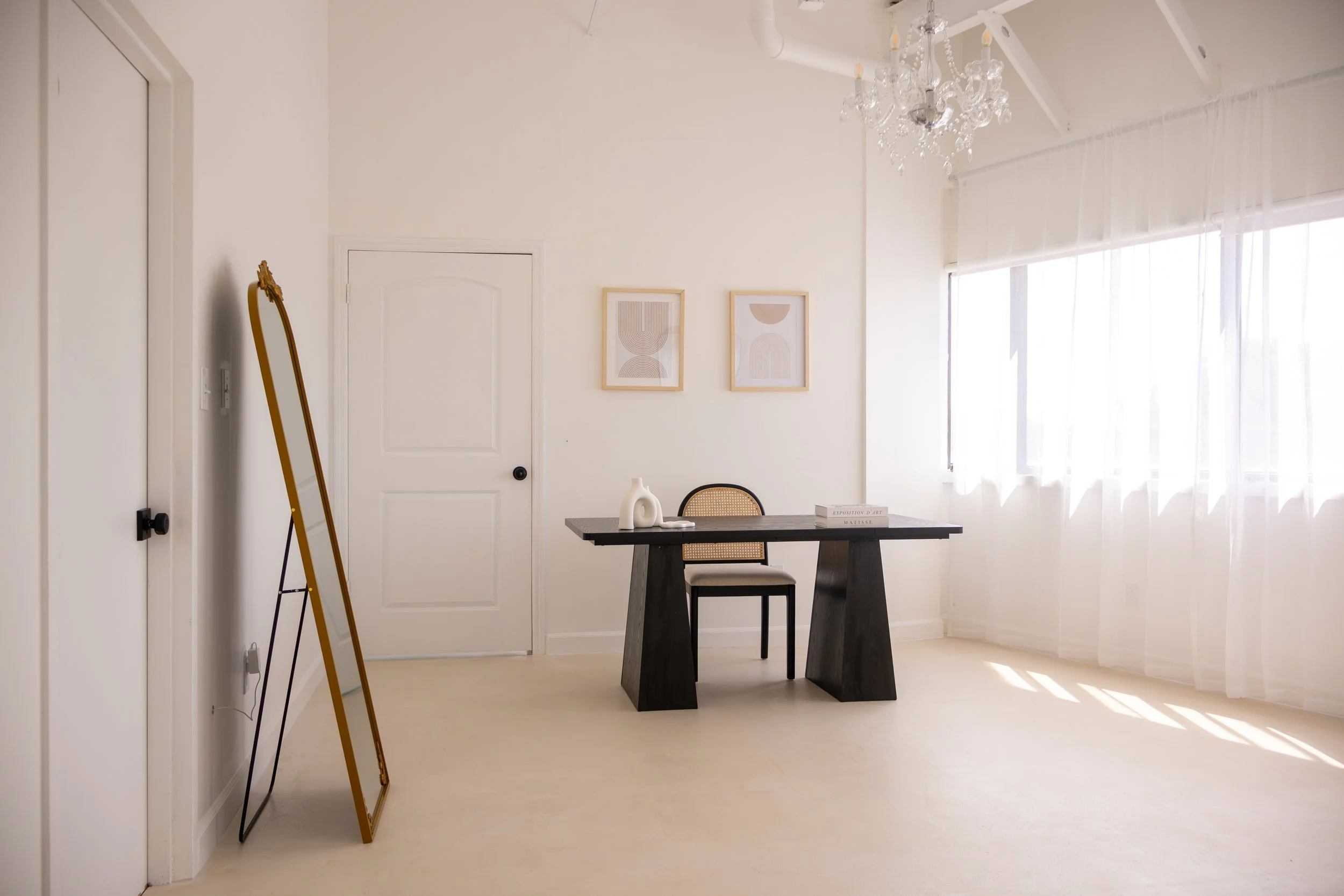 Minimalist dining room with a black table, a single chair, white walls, two framed abstract art pieces, a mirror leaning against the wall, and sheer white curtains letting in natural light.