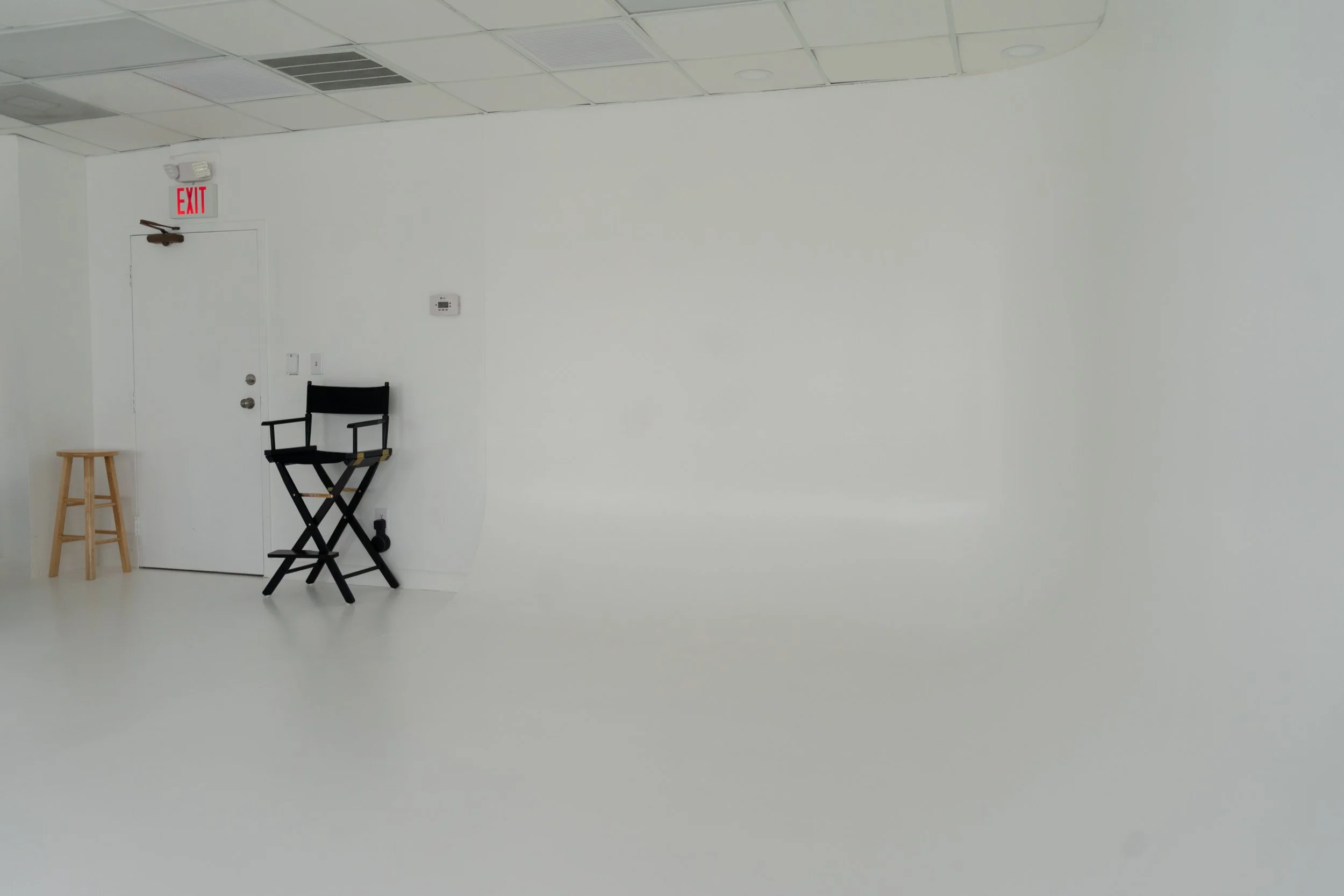 A minimal studio space with a white wall, ceiling, and floor. There is a door with an exit sign above it, a small wooden stool to the left, a black director's chair, and an electronic device on the wall.