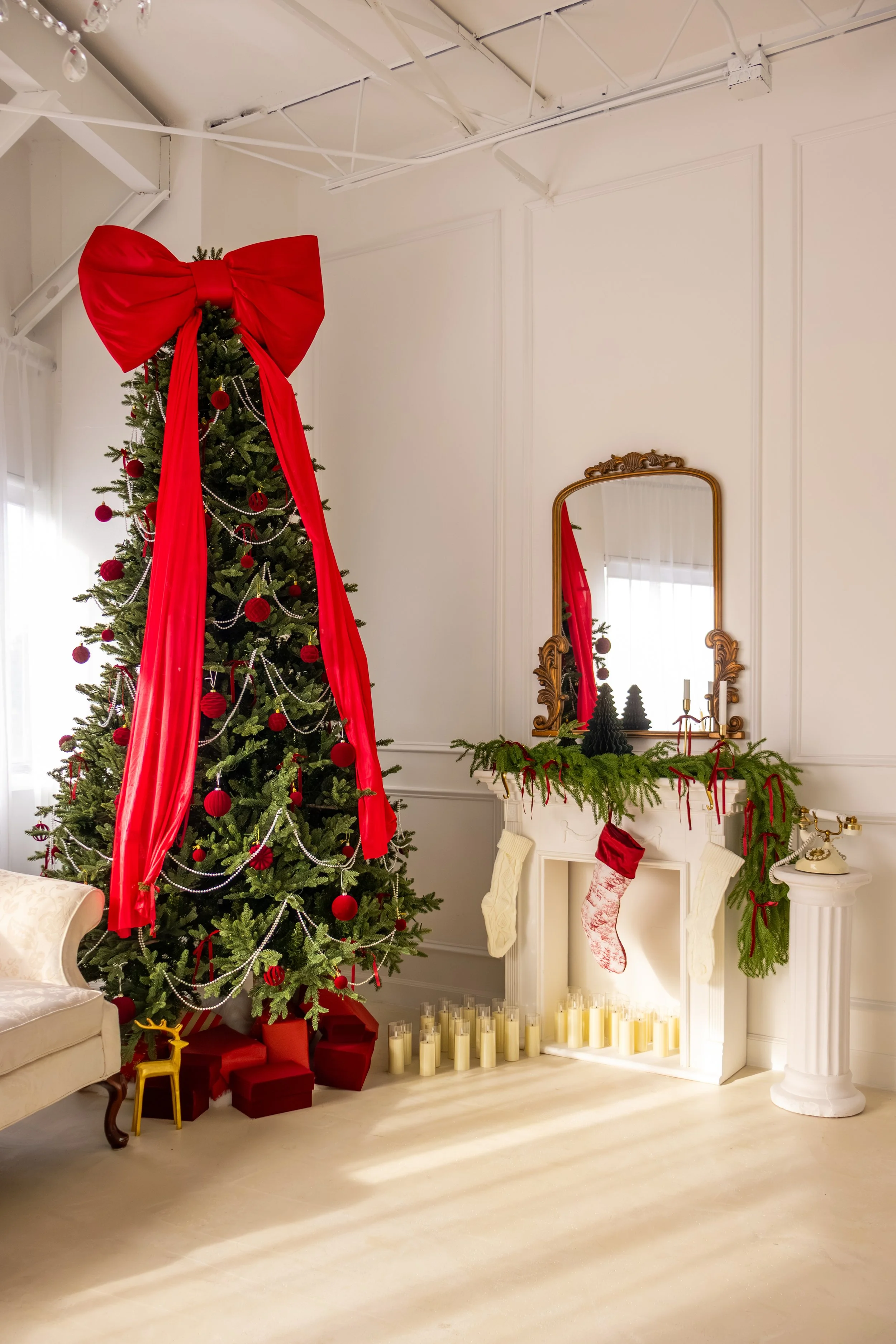 Decorated Christmas tree with red bow and ornaments next to a fireplace with stockings and candles in a bright room.